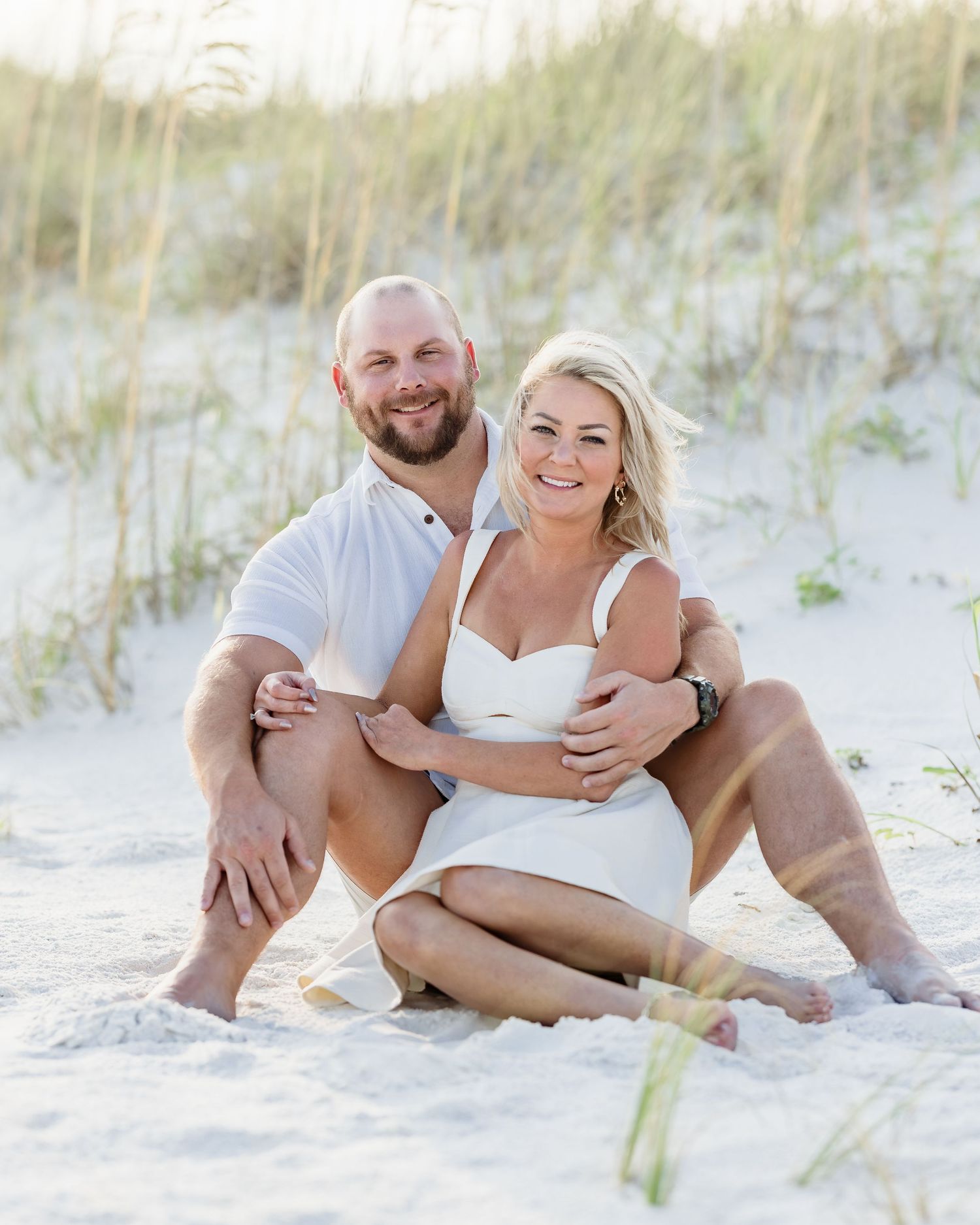 Happy couple sitting on sandy beach dunes, woman in white dress, man in white polo, smiling together outdoors.