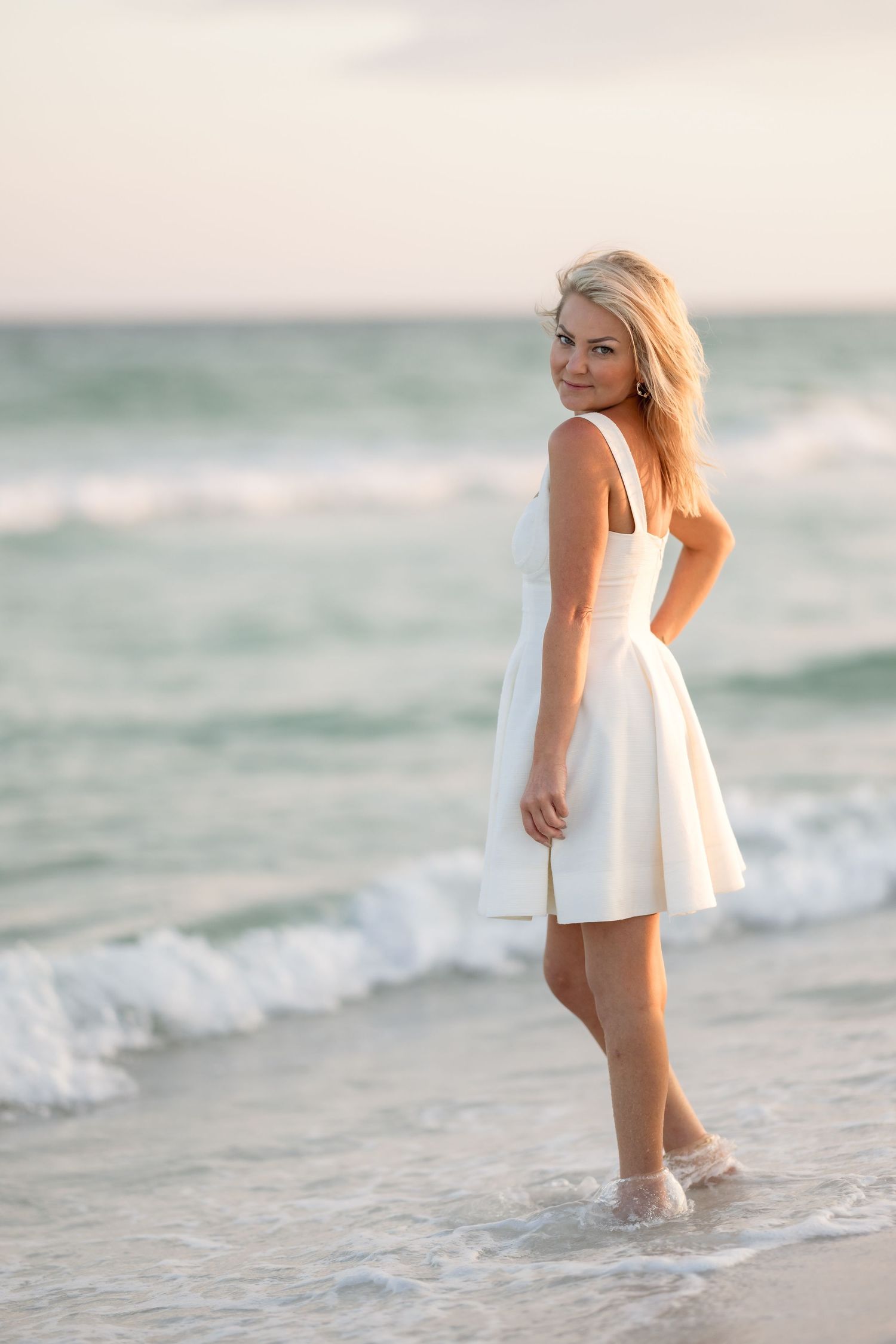 Blonde woman in white sundress standing in ocean waves on sandy beach at sunset.