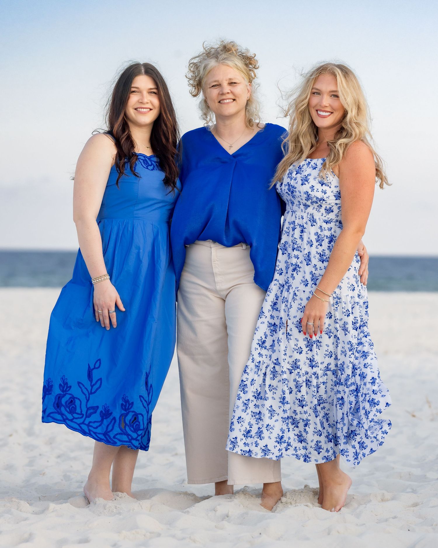 Three women in blue outfits smiling together on a sandy beach under a clear sky.
