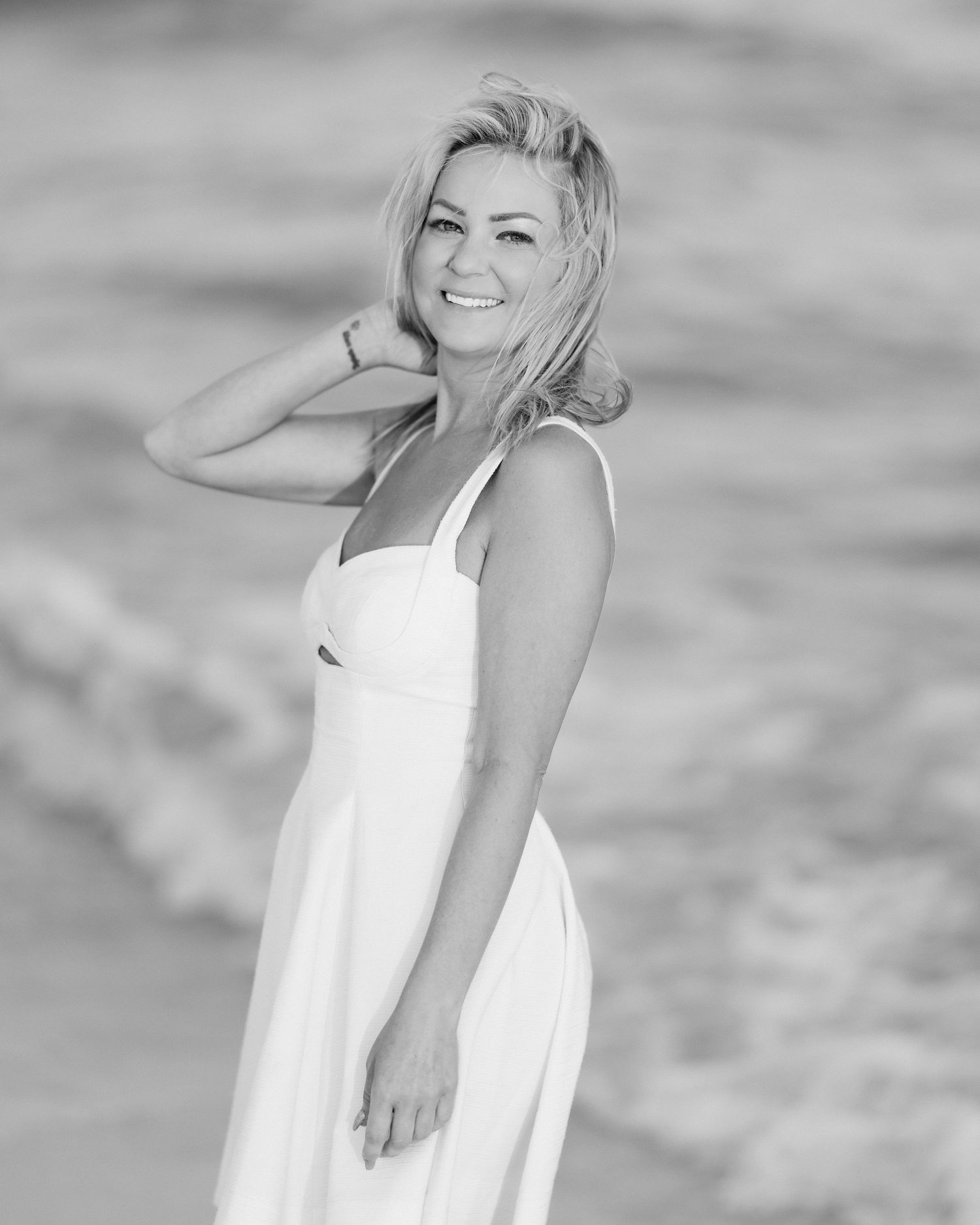 Black and white photo of smiling blonde woman in white dress posing near ocean waves.