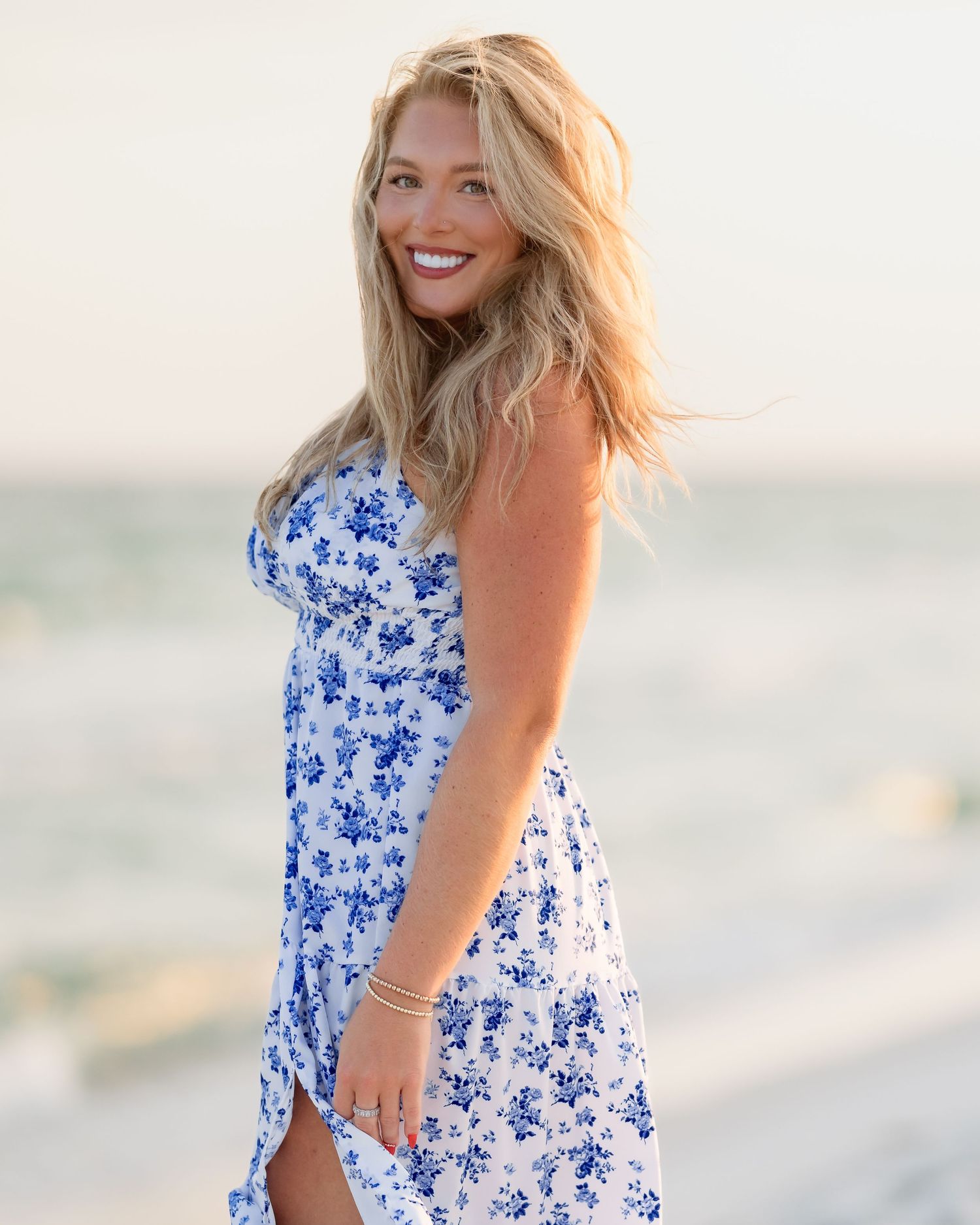 Blonde woman in blue floral dress smiles over her shoulder on a beach at sunset.