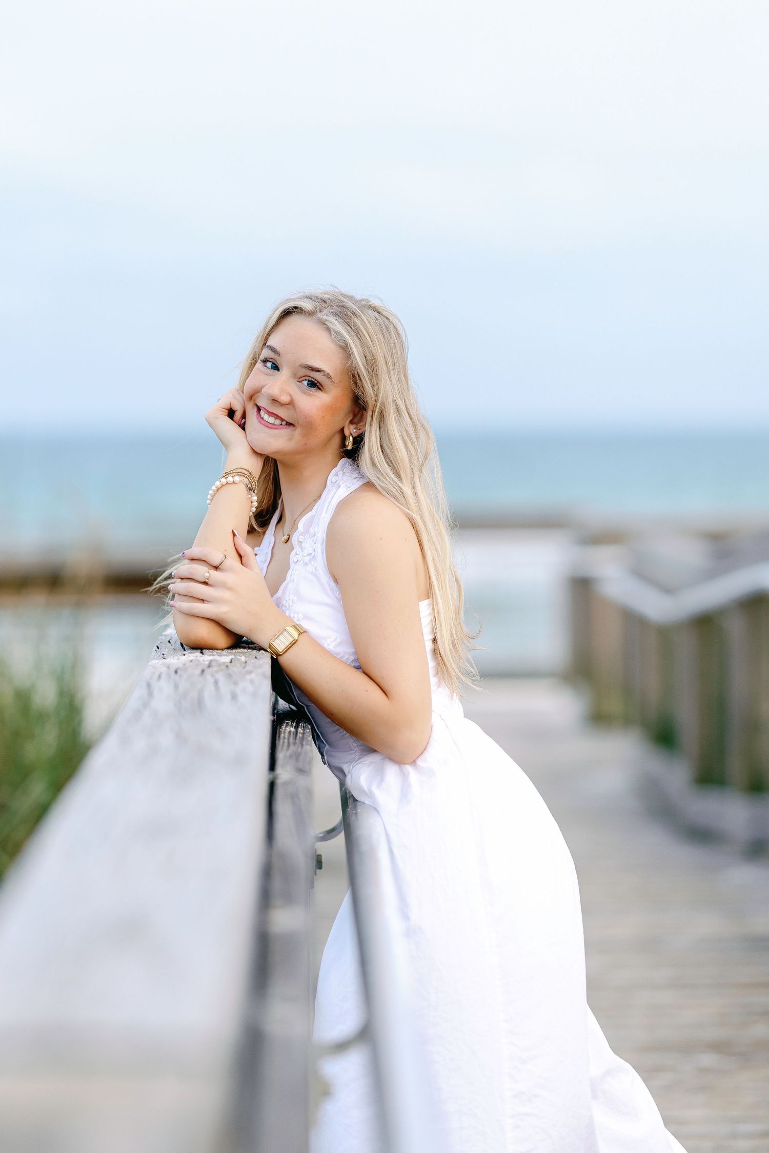 Contemplative blonde woman in white dress leans against wooden railing on beach boardwalk at sunset.