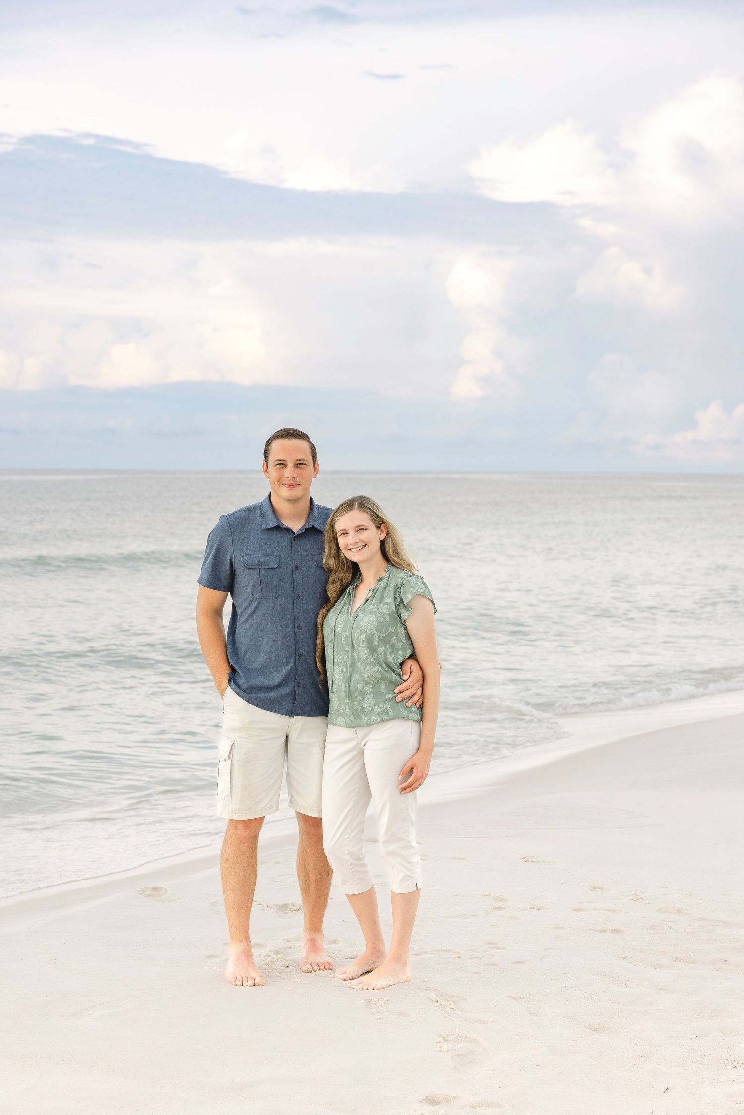 A couple stands together barefoot on a pristine white sandy beach at sunset.