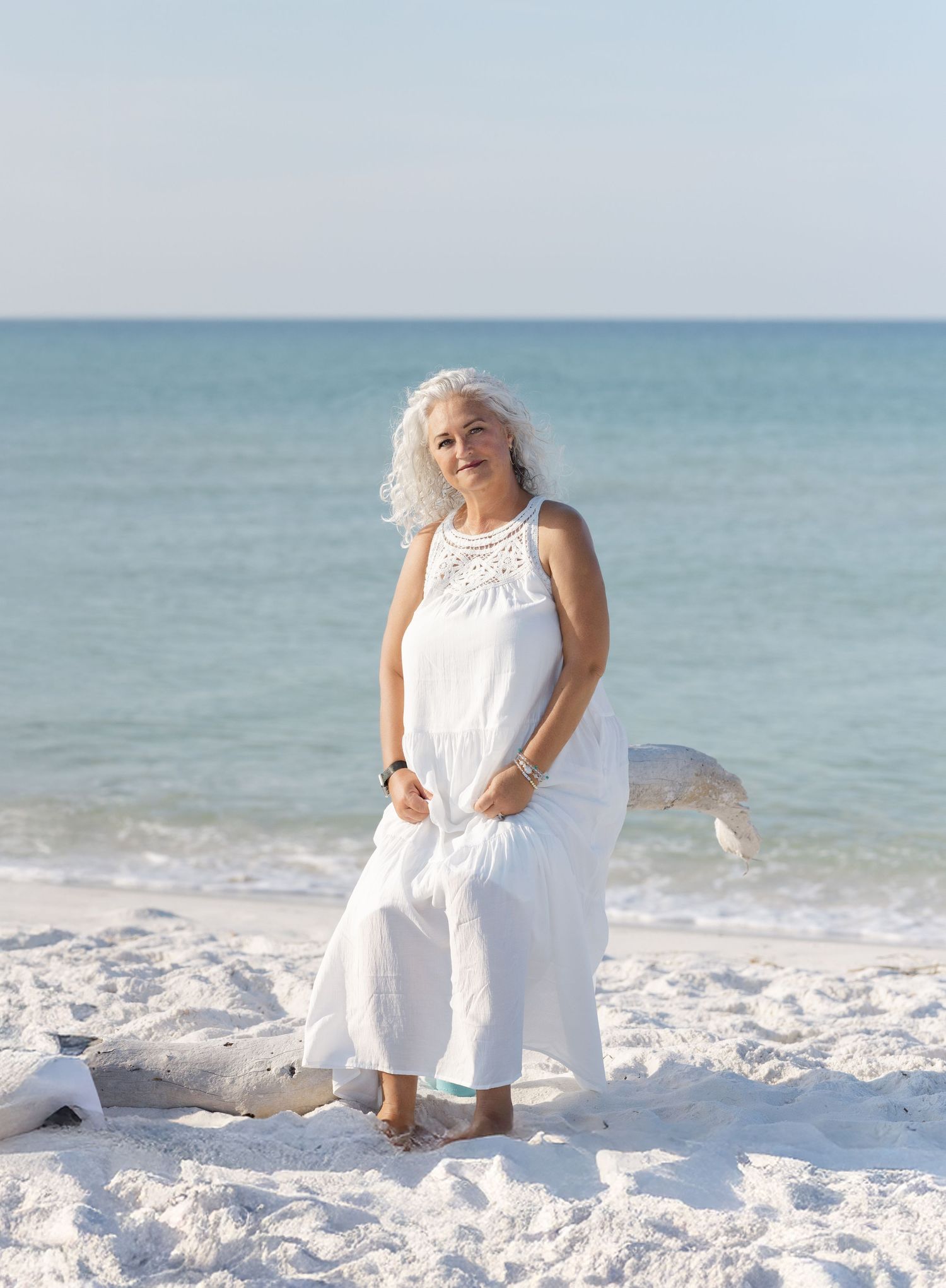 A person in a flowing white dress stands serenely on a pristine white sandy beach with turquoise ocean waters behind.