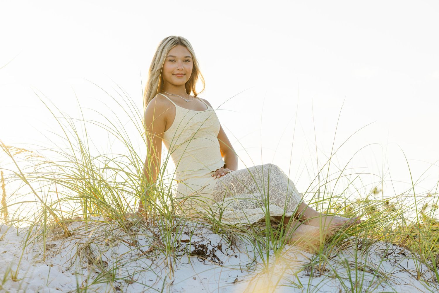A woman in a white dress poses gracefully on a sandy beach dune with tall grass during golden hour.