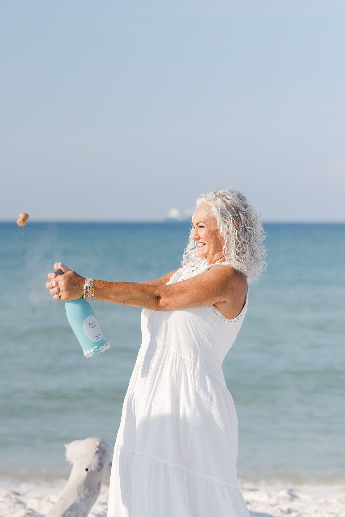 Person in flowing white dress sprays a water bottle while standing on a sunny beach with blue ocean in the background.