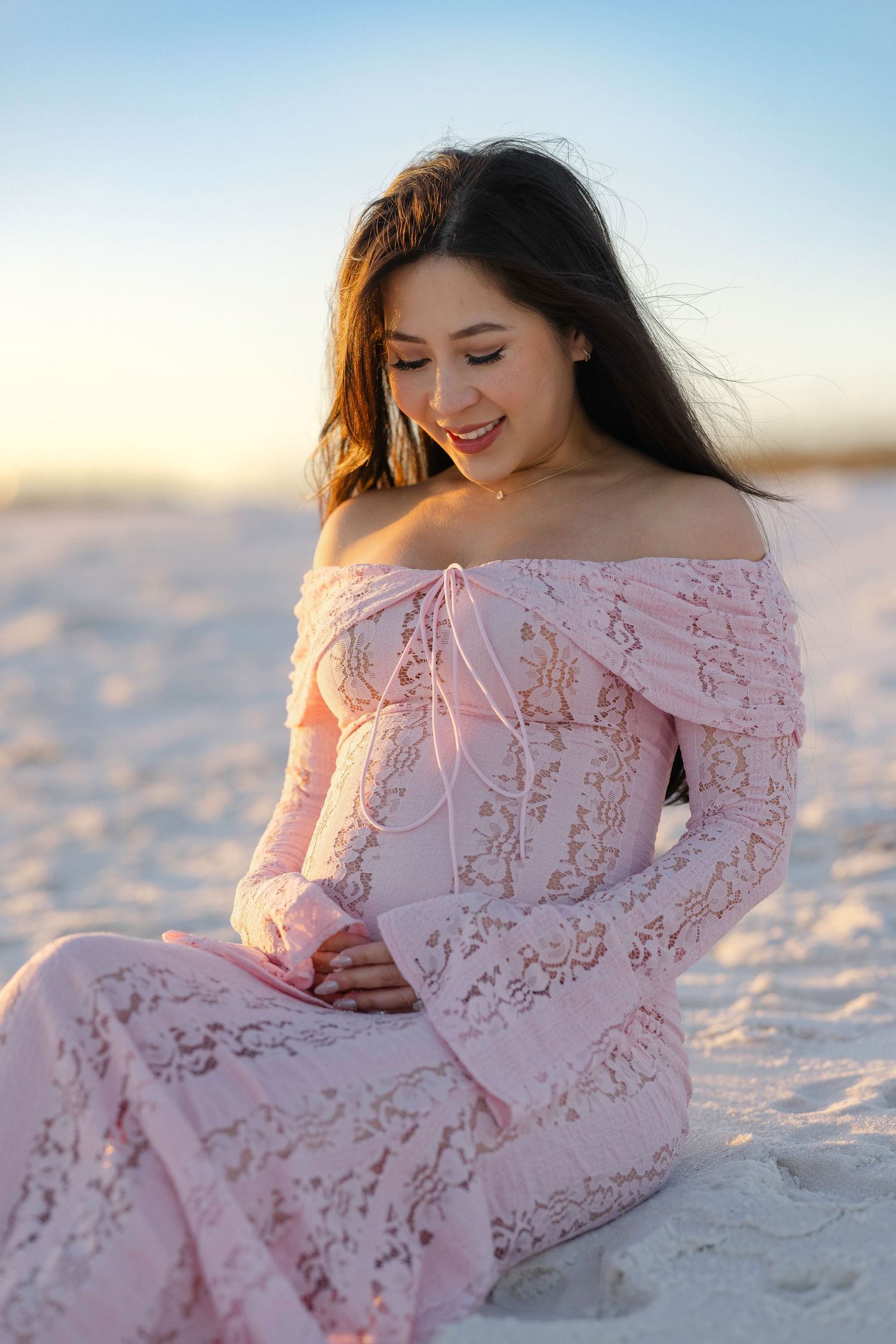 A person in a pink lace dress reclines on pristine white sand during a beach photoshoot at golden hour.