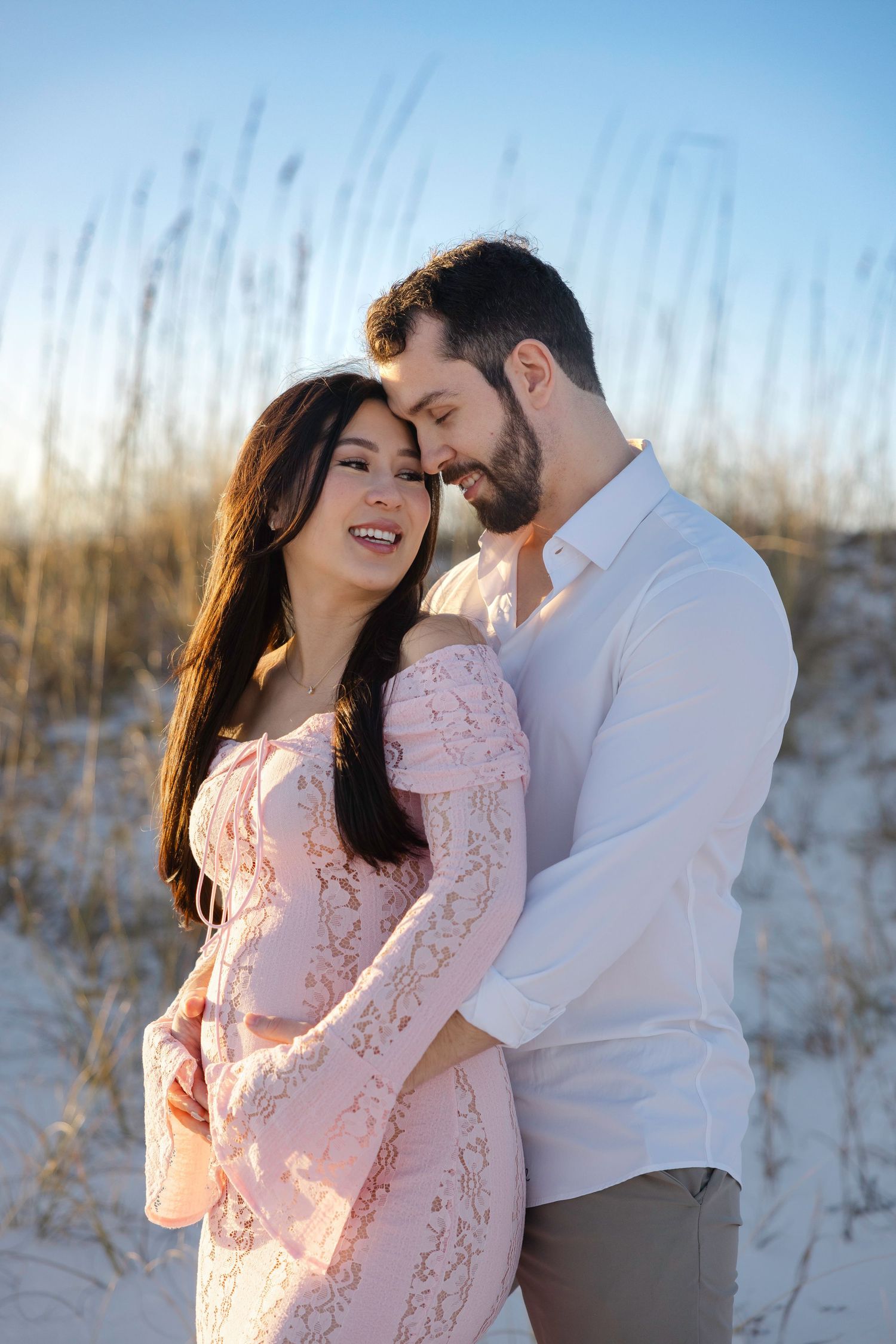A couple embraces on a sandy beach with sea oats and soft lighting during a romantic photoshoot.