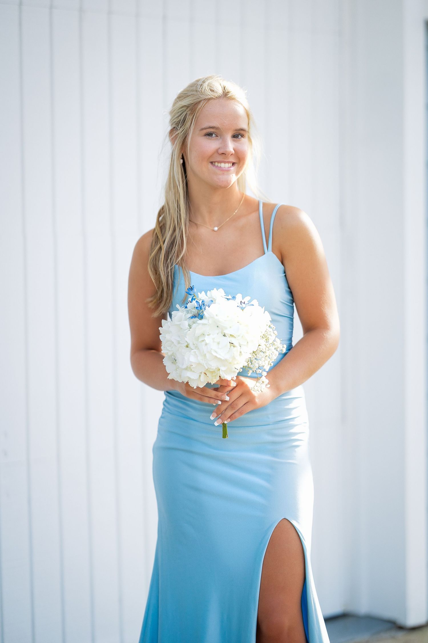 A woman in a light blue formal dress holds a white floral bouquet against a white backdrop.