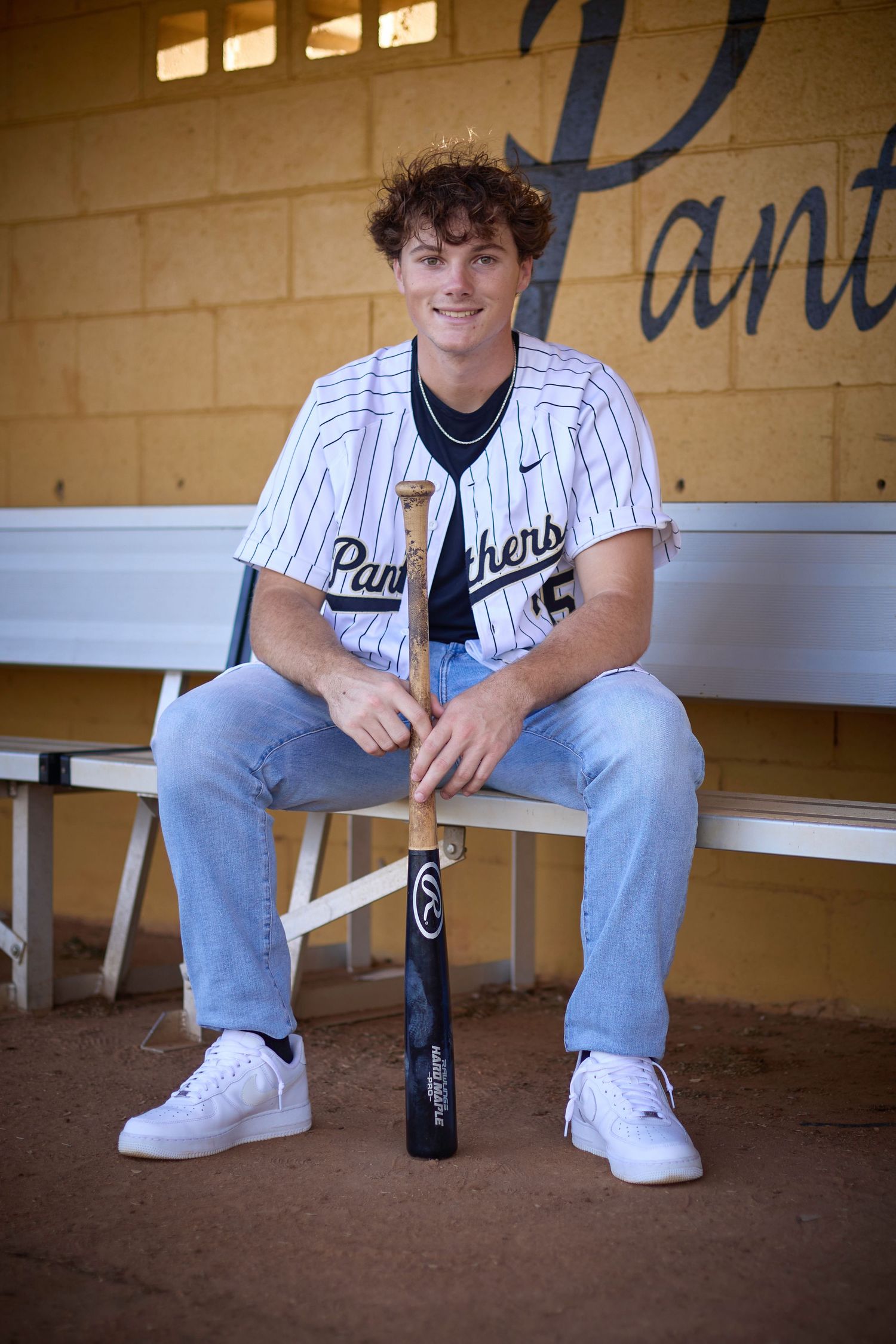 Baseball player in pinstripe uniform and jeans sits in dugout with bat while wearing white sneakers.