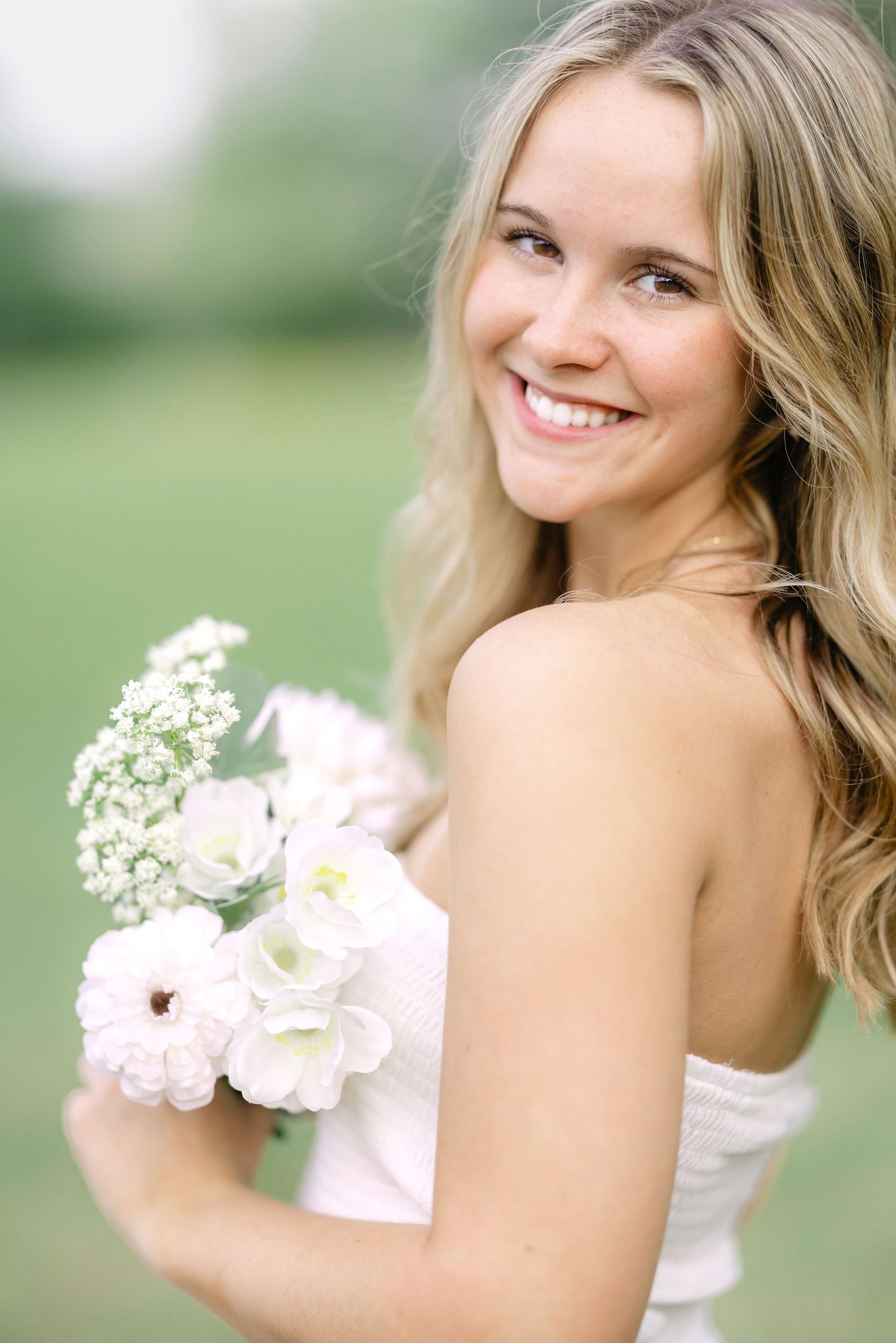 A bride in a white dress holds a delicate bouquet of white flowers against a soft outdoor background.