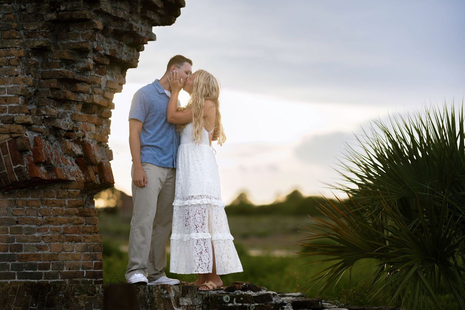 Couple sharing a romantic kiss near rustic brick ruins at sunset, woman in white dress, man in blue shirt.