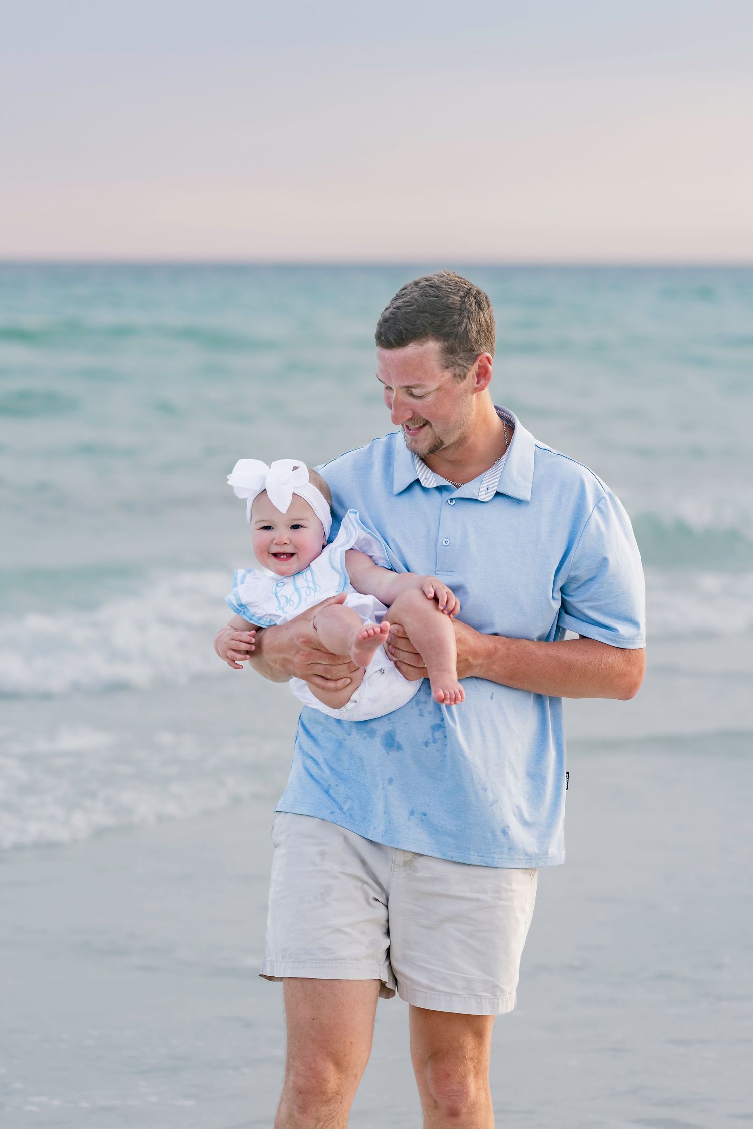 Smiling man in blue shirt holds a baby girl with a white bow while standing at the ocean's edge.