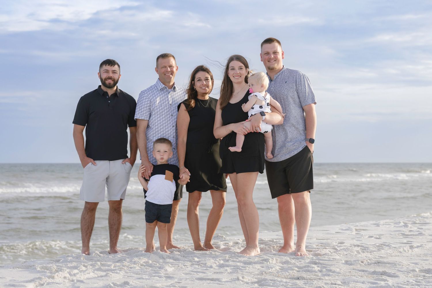 Family of seven posing together on a sandy beach with ocean waves in the background on a cloudy day.