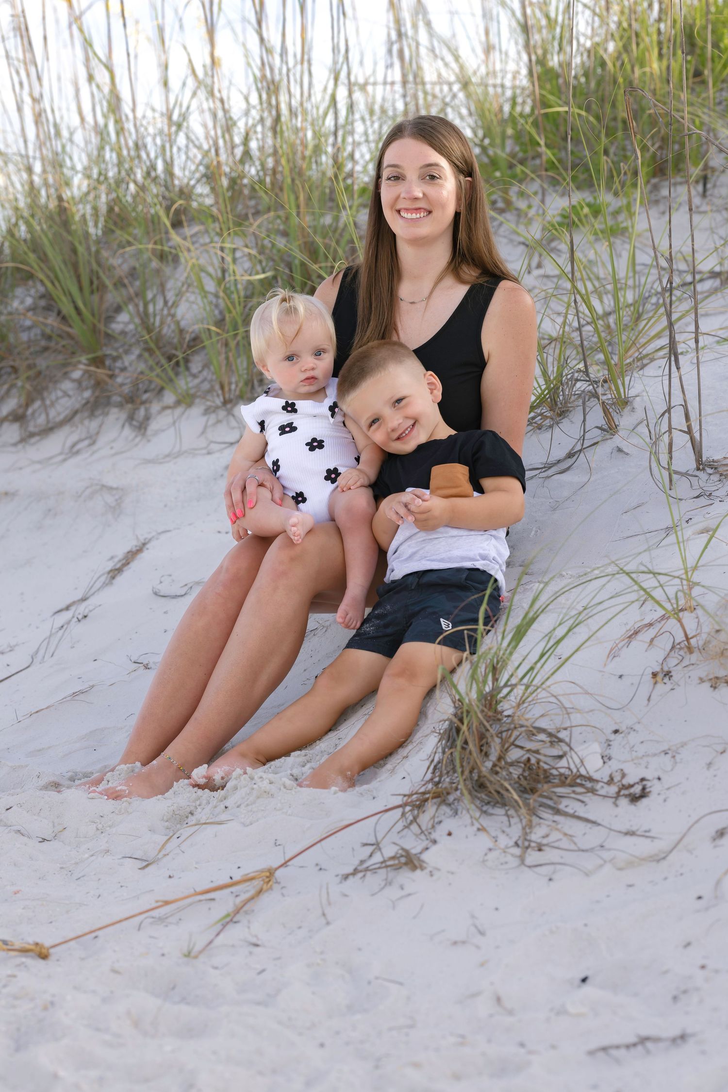Young woman sits on beach sand dunes with two small children in her lap, smiling at camera.
