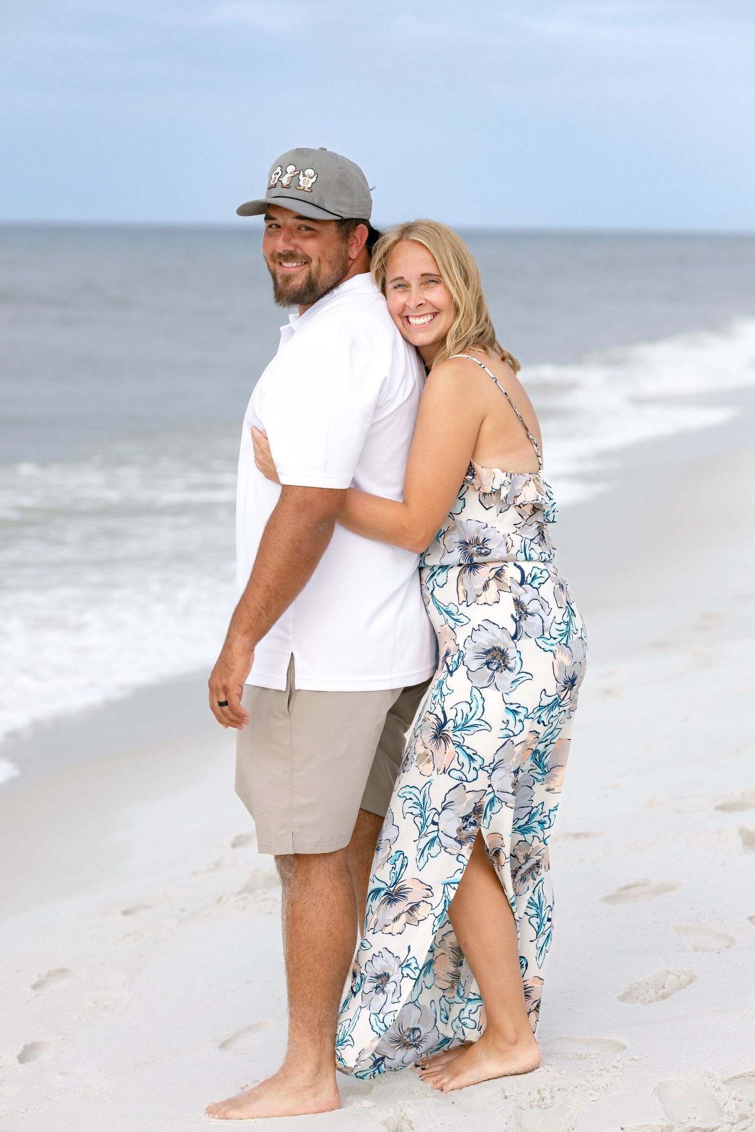 Couple embracing on sunny beach, woman in floral maxi dress hugging man in white shirt and cap.