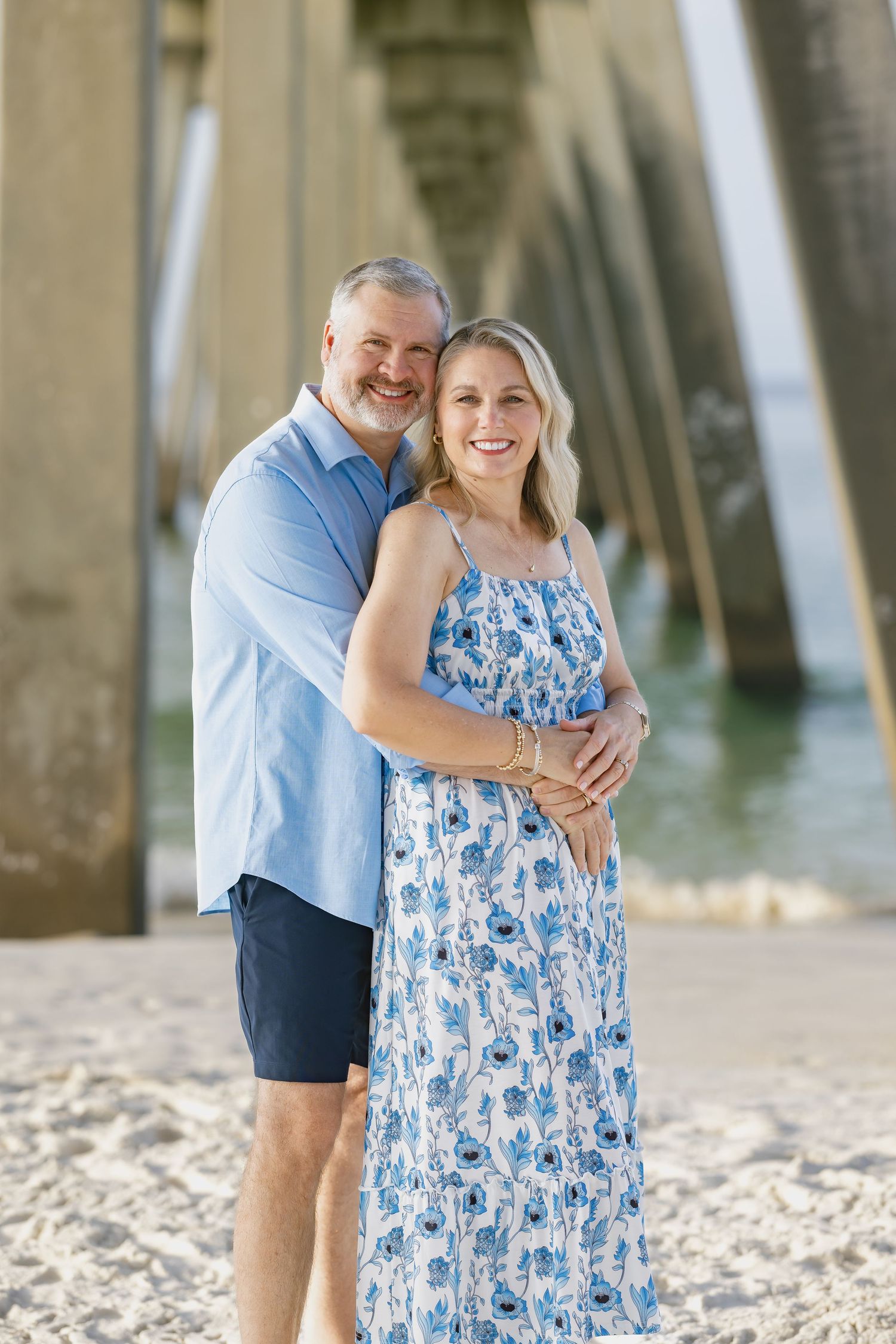 Smiling couple embracing on a sandy beach under a pier, wearing coordinating blue outfits.