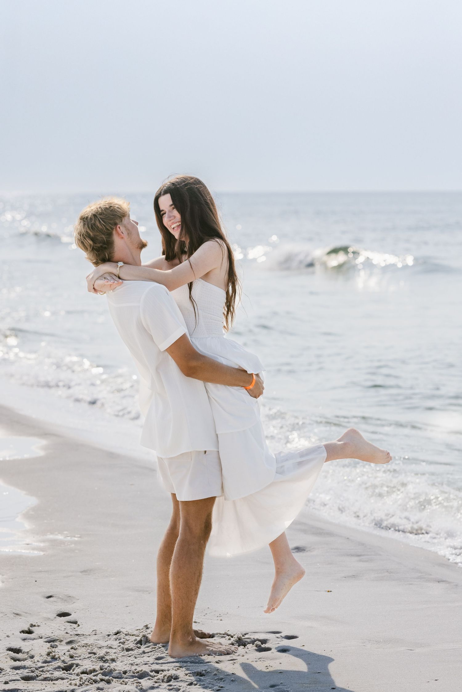 Man lifting woman in white dress on a sandy beach with ocean waves in the background.