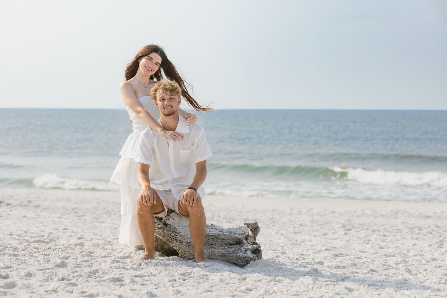 Couple posing on driftwood at a white sand beach, woman in white dress standing behind seated man, ocean in background.