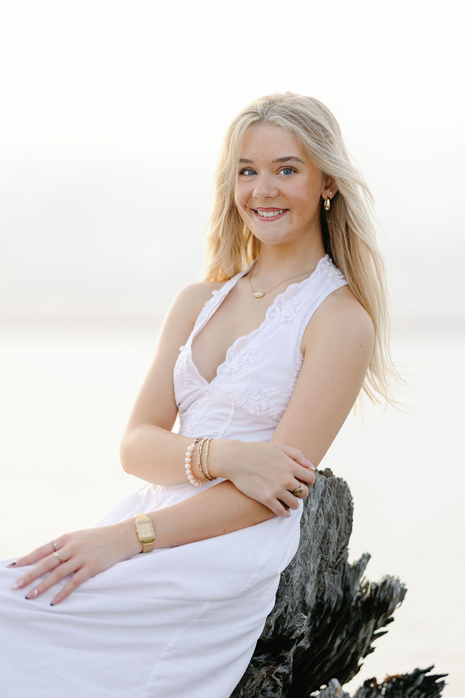 Smiling blonde woman in white lace dress sitting on driftwood at the beach wearing gold jewelry.