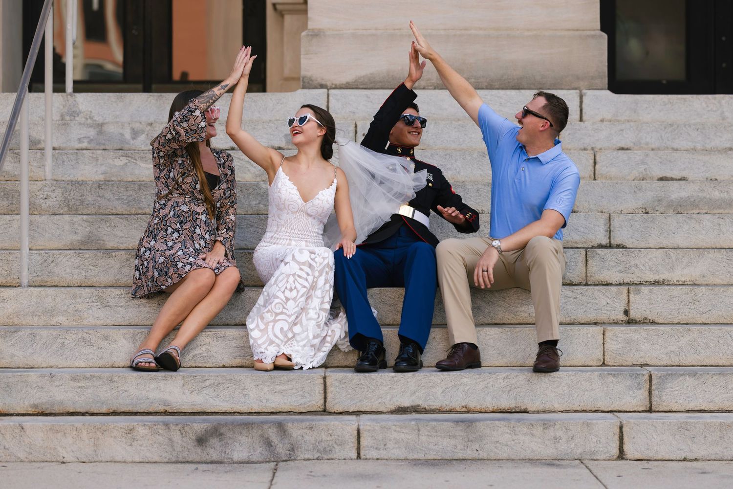 Four friends sitting on stone steps giving high fives in a chain, dressed in summer attire outdoors.