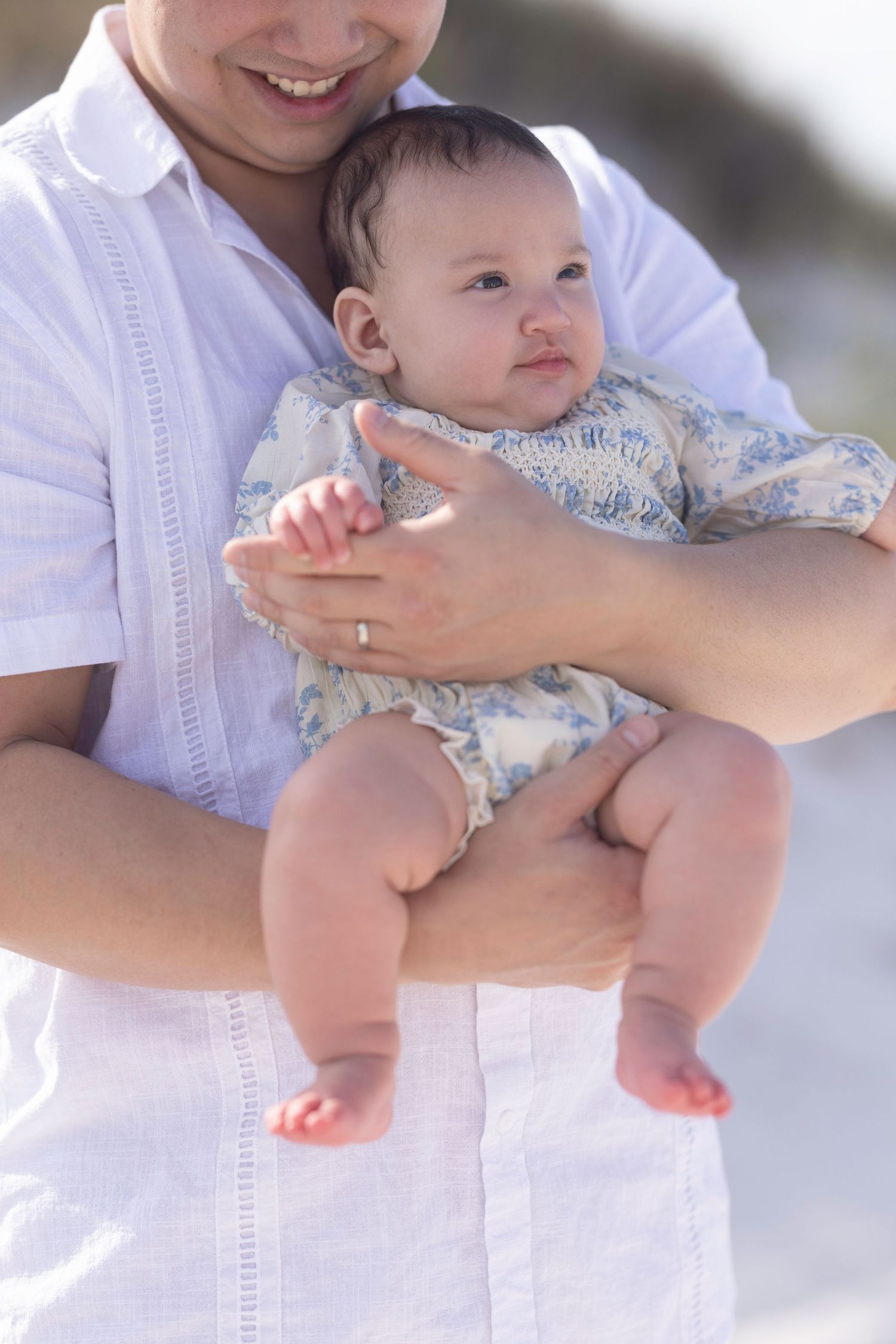 Father in white shirt holds infant baby at beach, smiling warmly while cradling the child in both arms outdoors.