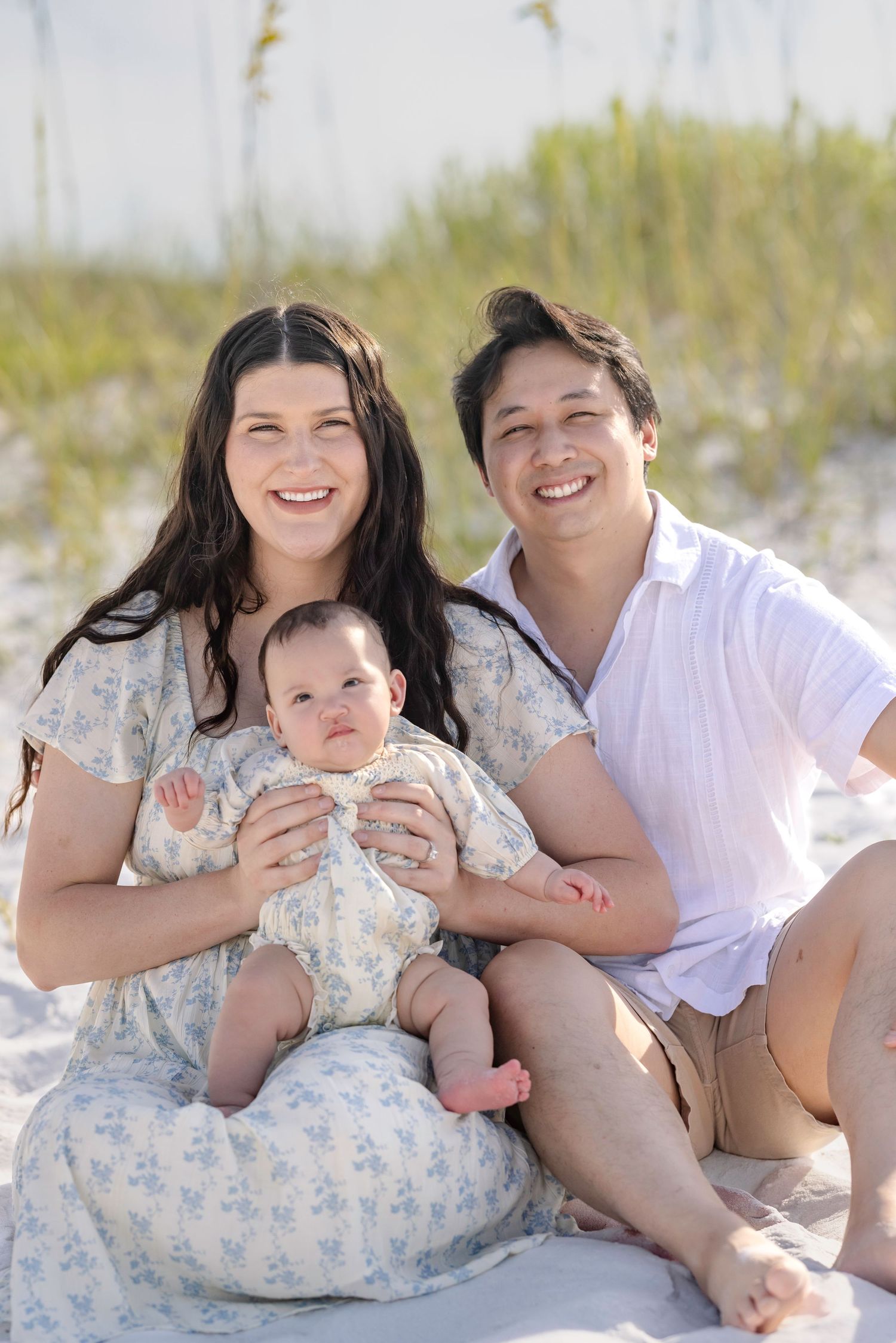 A young family smiles together on a sandy beach, with a mother and father holding their happy baby outdoors.