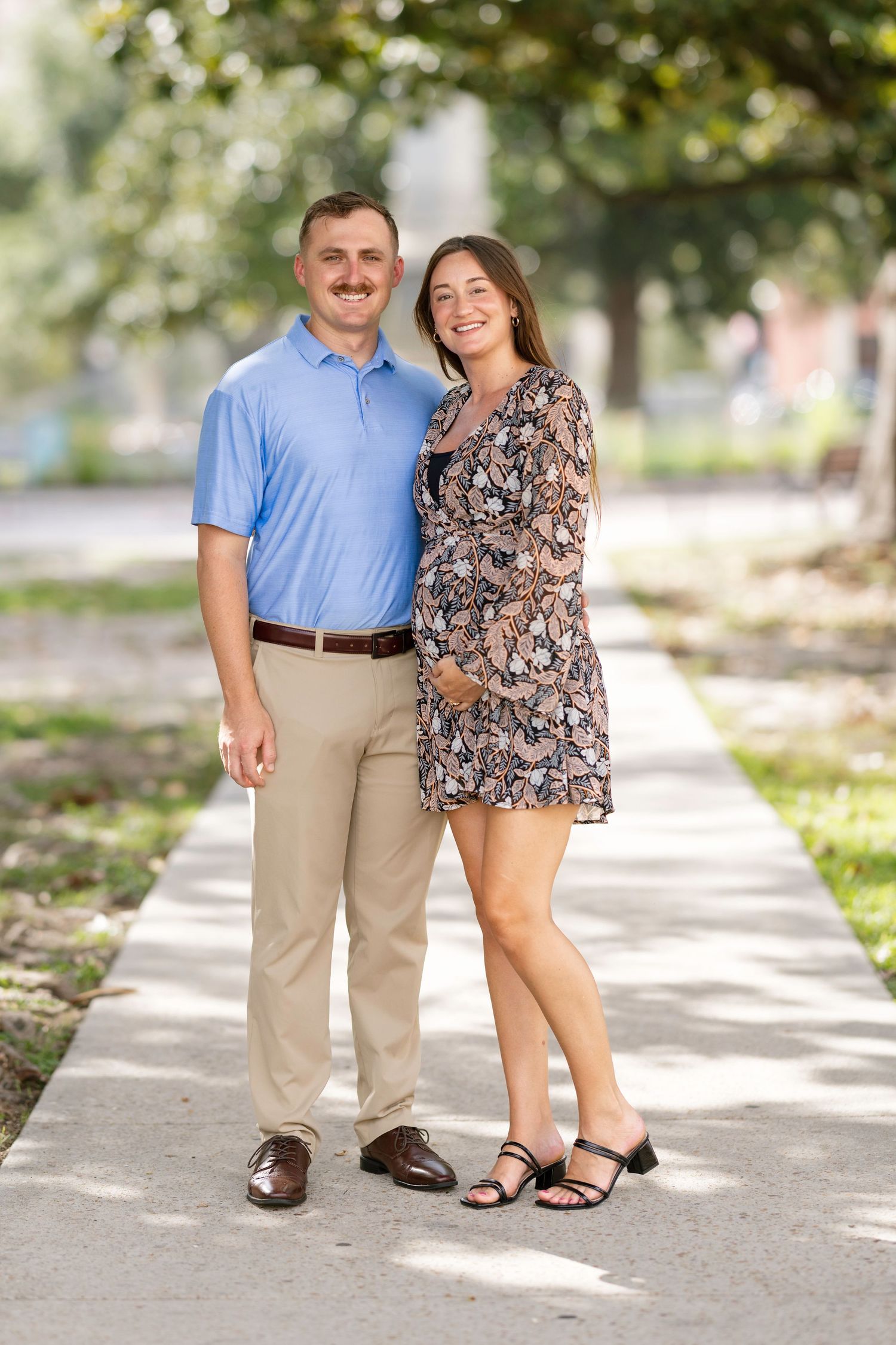 A smiling couple poses together on a sunny outdoor walkway, man in blue polo and khakis, woman in floral dress.