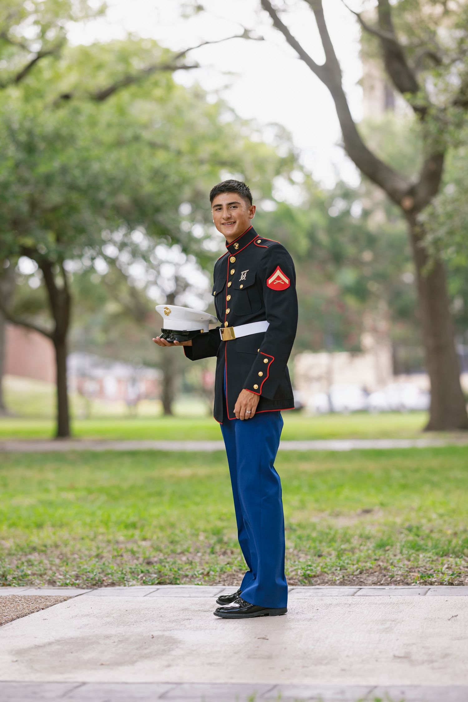 Young US Marine in dress blues uniform holding white cap, smiling outdoors in park setting.