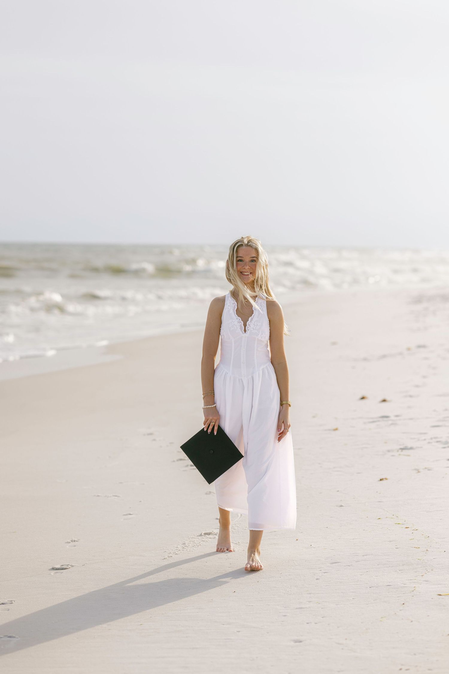 Young blonde woman in white maxi dress walks barefoot on sandy beach holding a dark clutch purse.