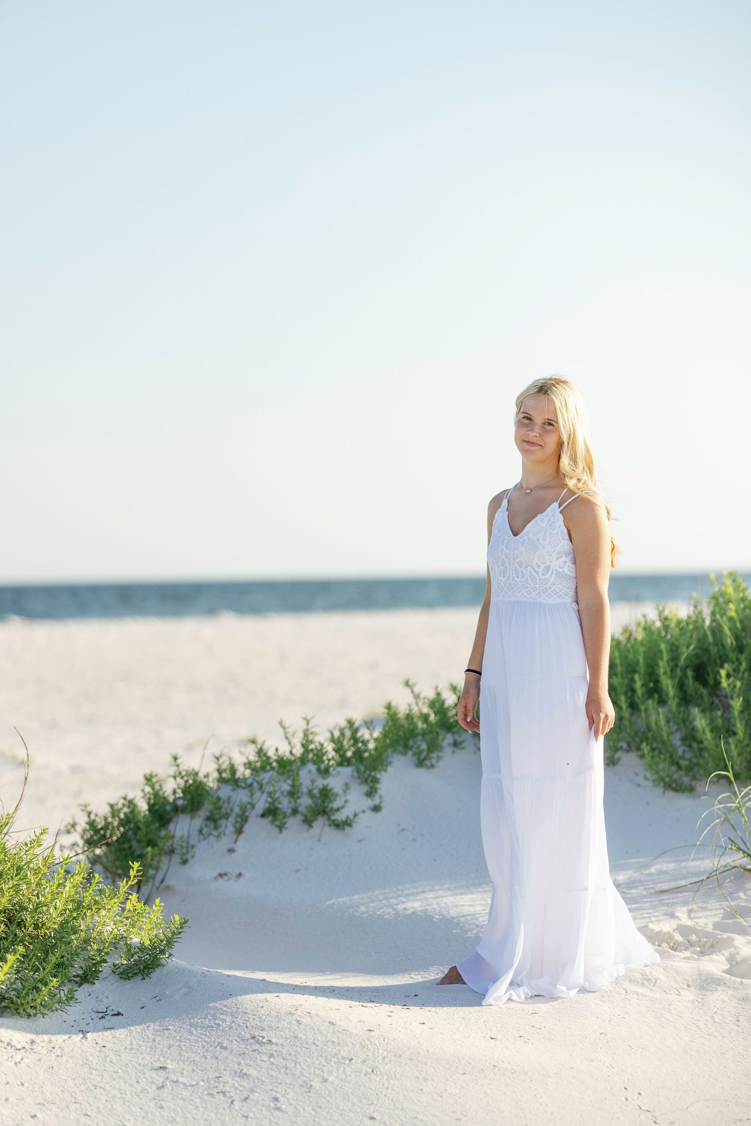 Young blonde woman in white maxi dress standing on white sand beach with ocean in background.