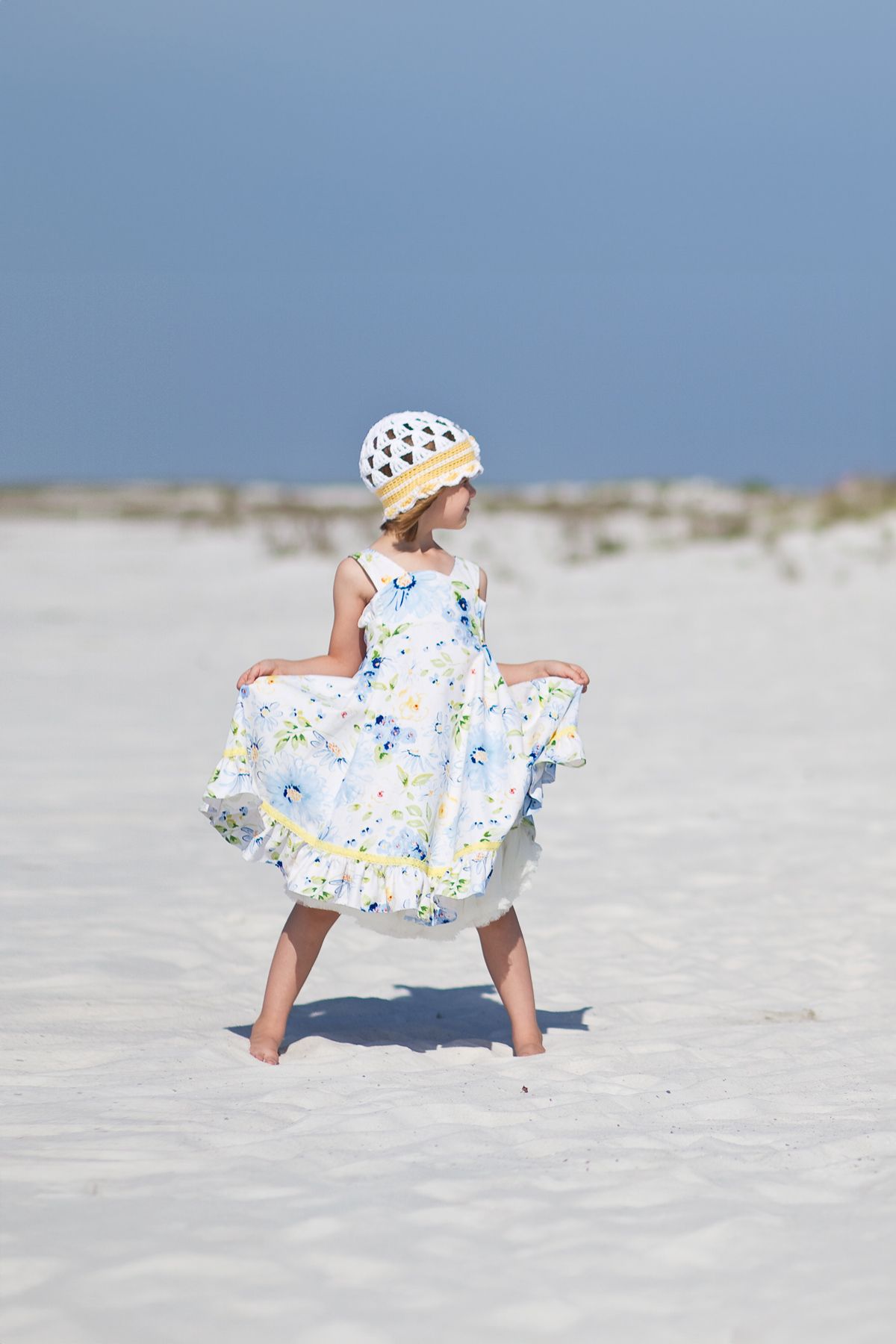 Young girl in floral dress and sun hat twirling on white sand beach with blue sky background.