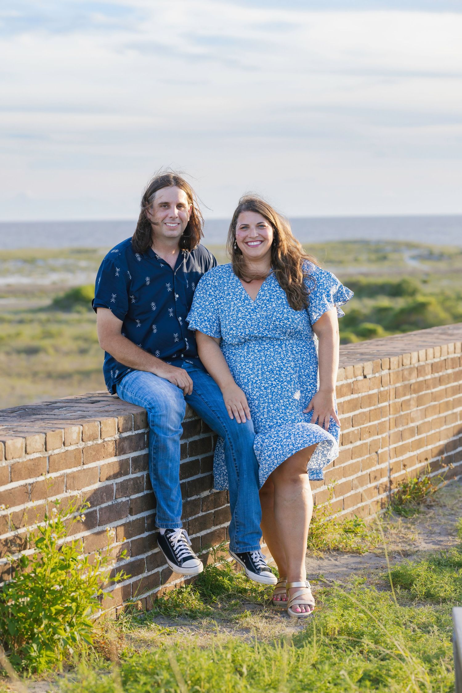 Smiling couple sitting on a brick wall with coastal scenery in the background on a sunny day.