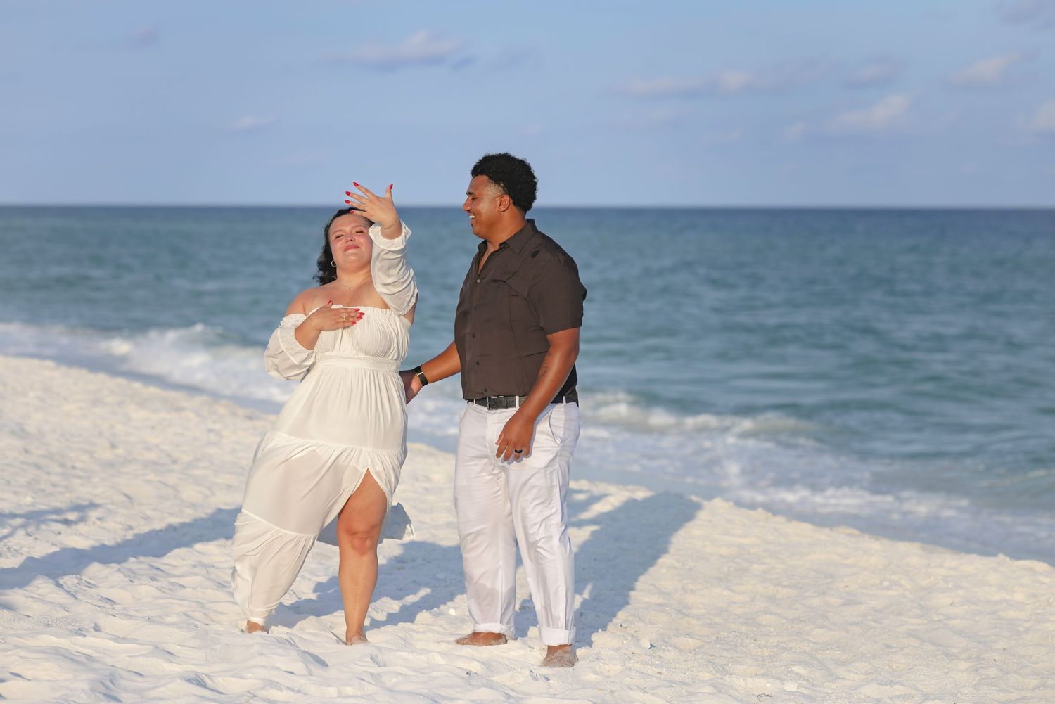 Couple in white outfits enjoying a romantic beach walk on white sand with calm blue ocean waves in the background.