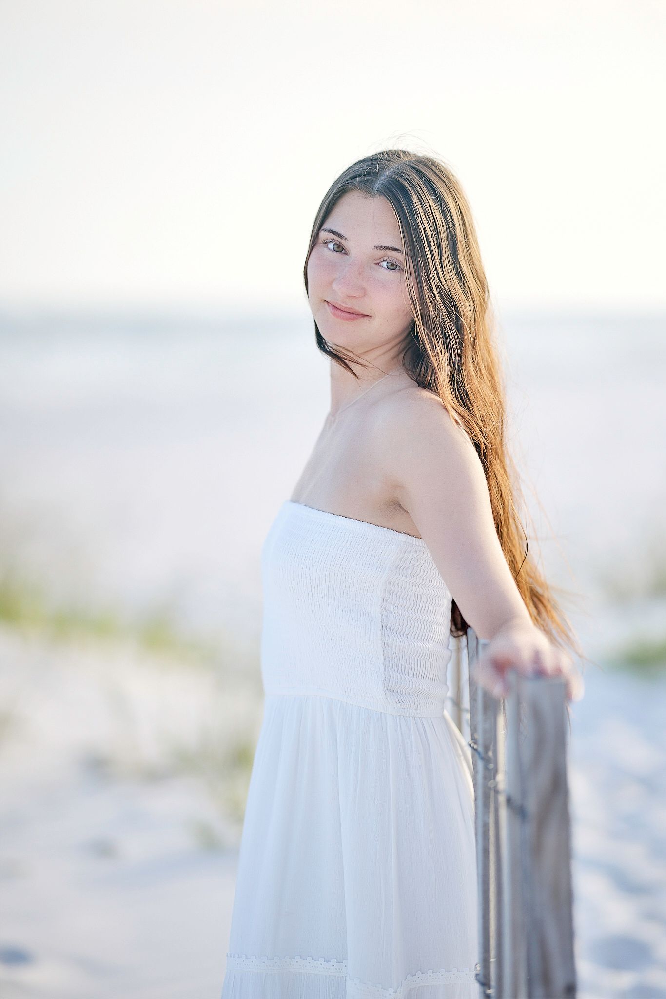Young woman in white strapless dress standing on sandy beach, leaning on wooden post with gentle backlight.