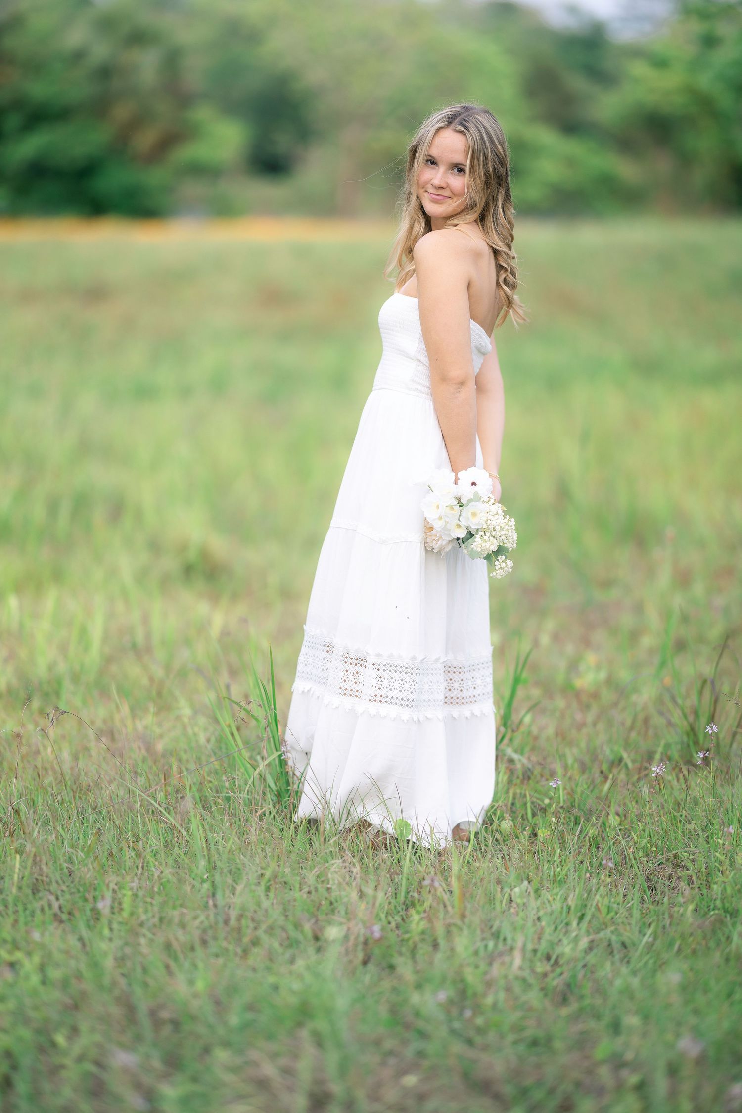 Elegant blonde woman in white strapless maxi dress holding bouquet while standing gracefully in green meadow.