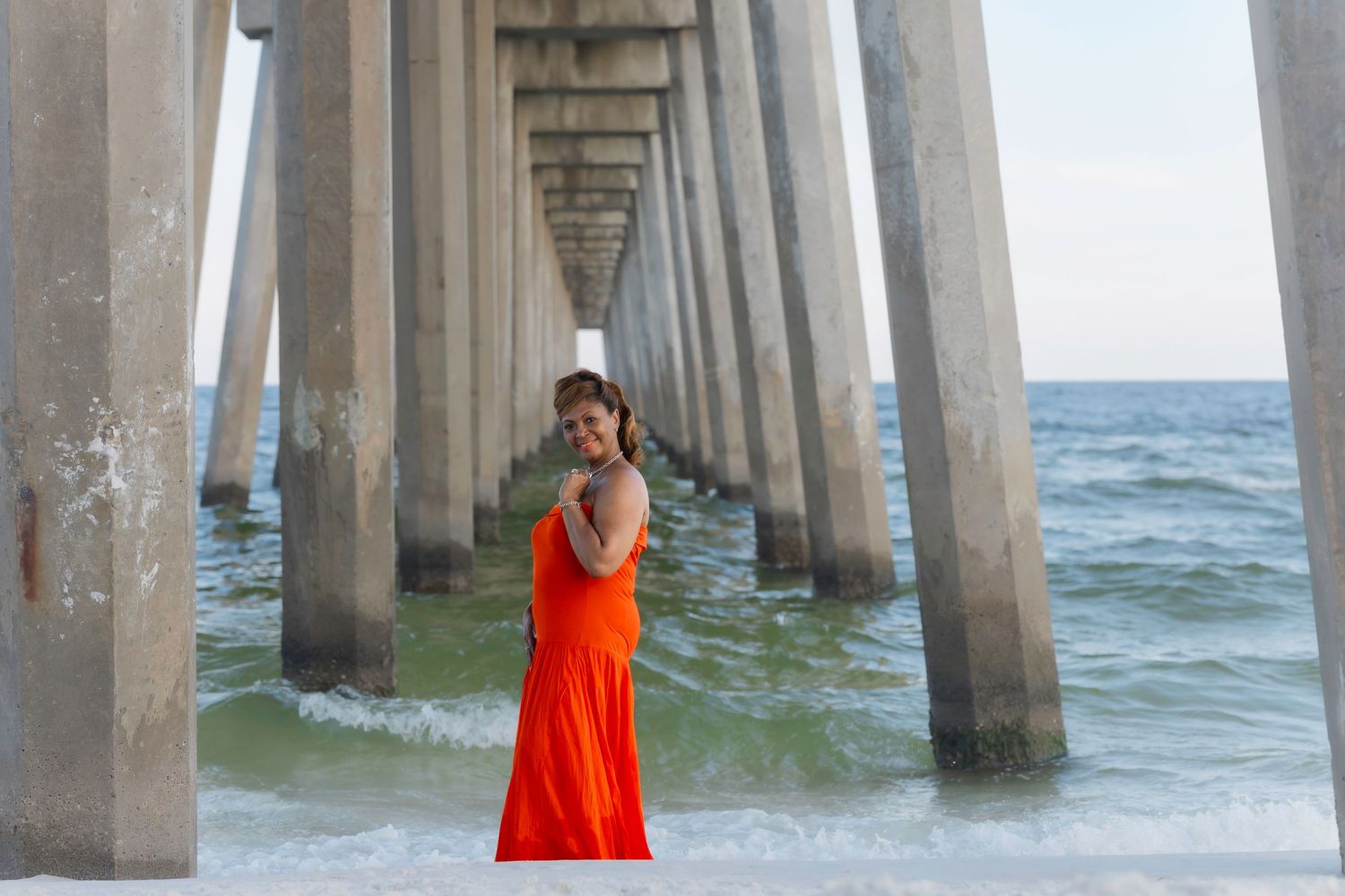 A woman in an orange dress poses gracefully under a beach pier with ocean waves behind her.