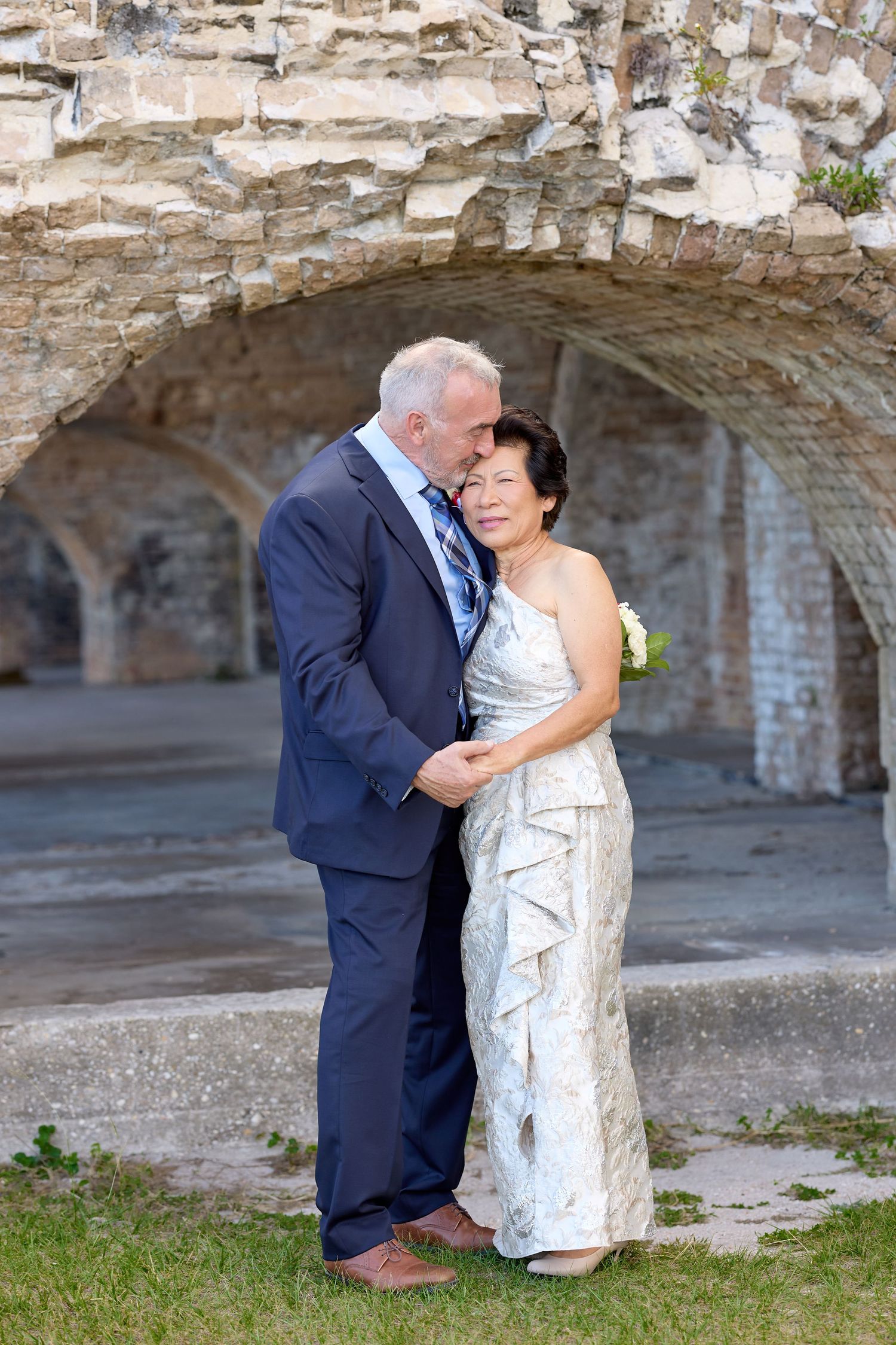 Elegant couple in wedding attire embracing before stone arched ruins, woman in white gown, man in navy suit.