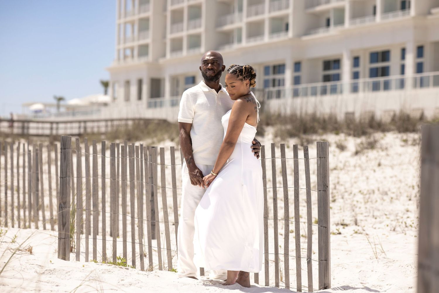 A couple in white attire poses romantically by a beach fence with a resort building in the background.