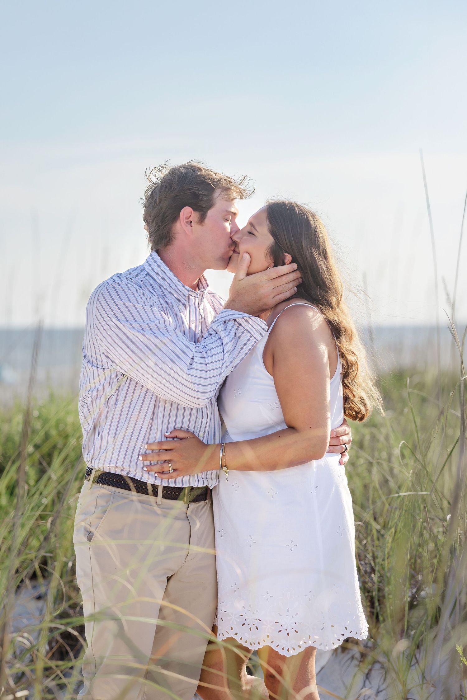 Couple sharing a romantic kiss on a sandy beach with sea grass and ocean in the background.