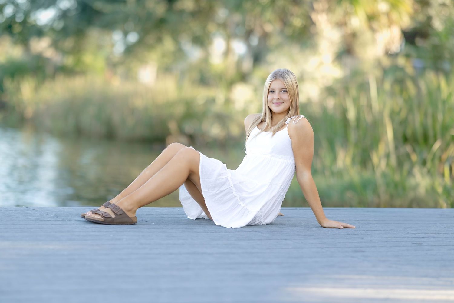 Young blonde woman in white dress sitting on wooden dock by serene lake surrounded by lush green trees.