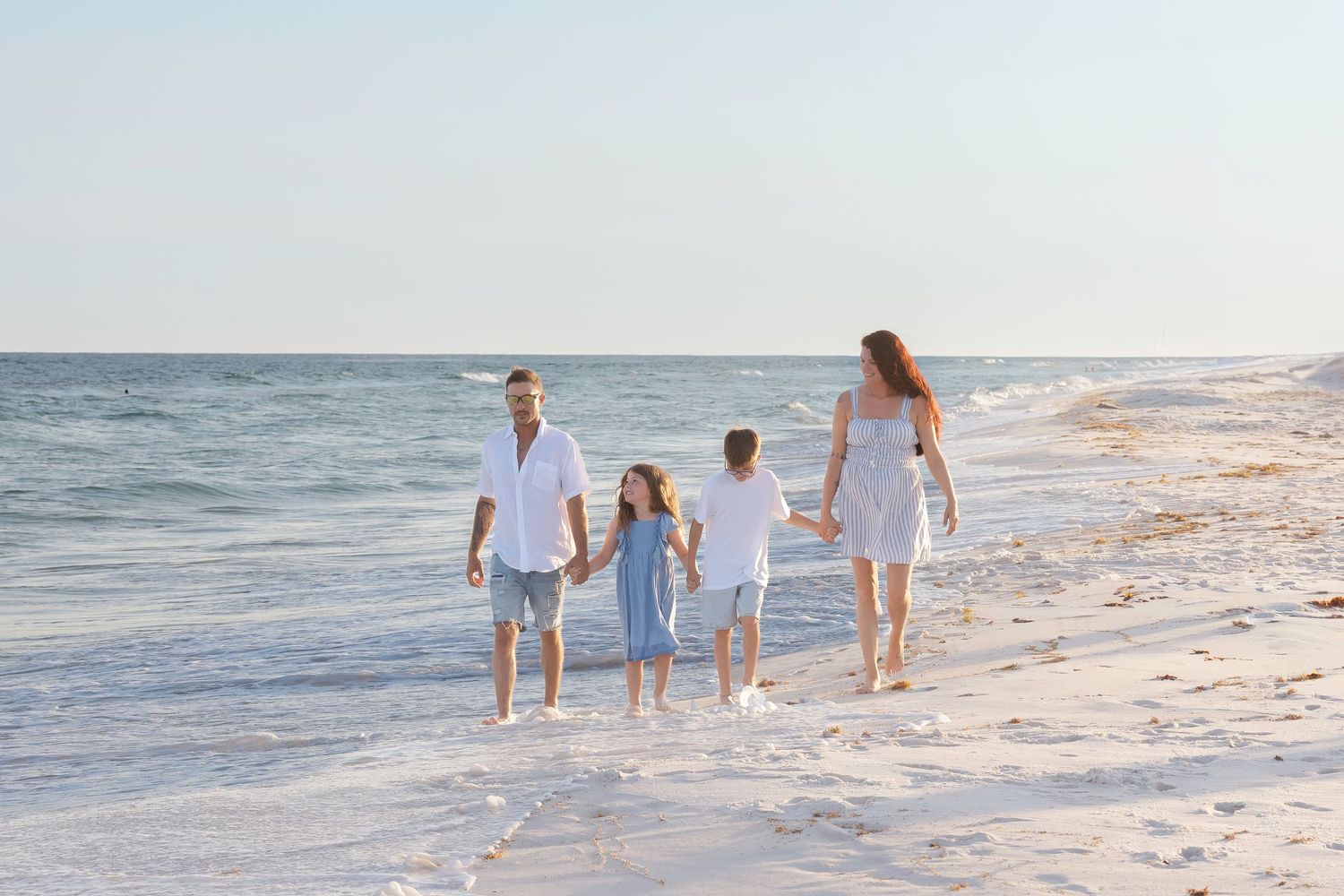 Family of four walking hand-in-hand along a sunny white sand beach near the ocean.