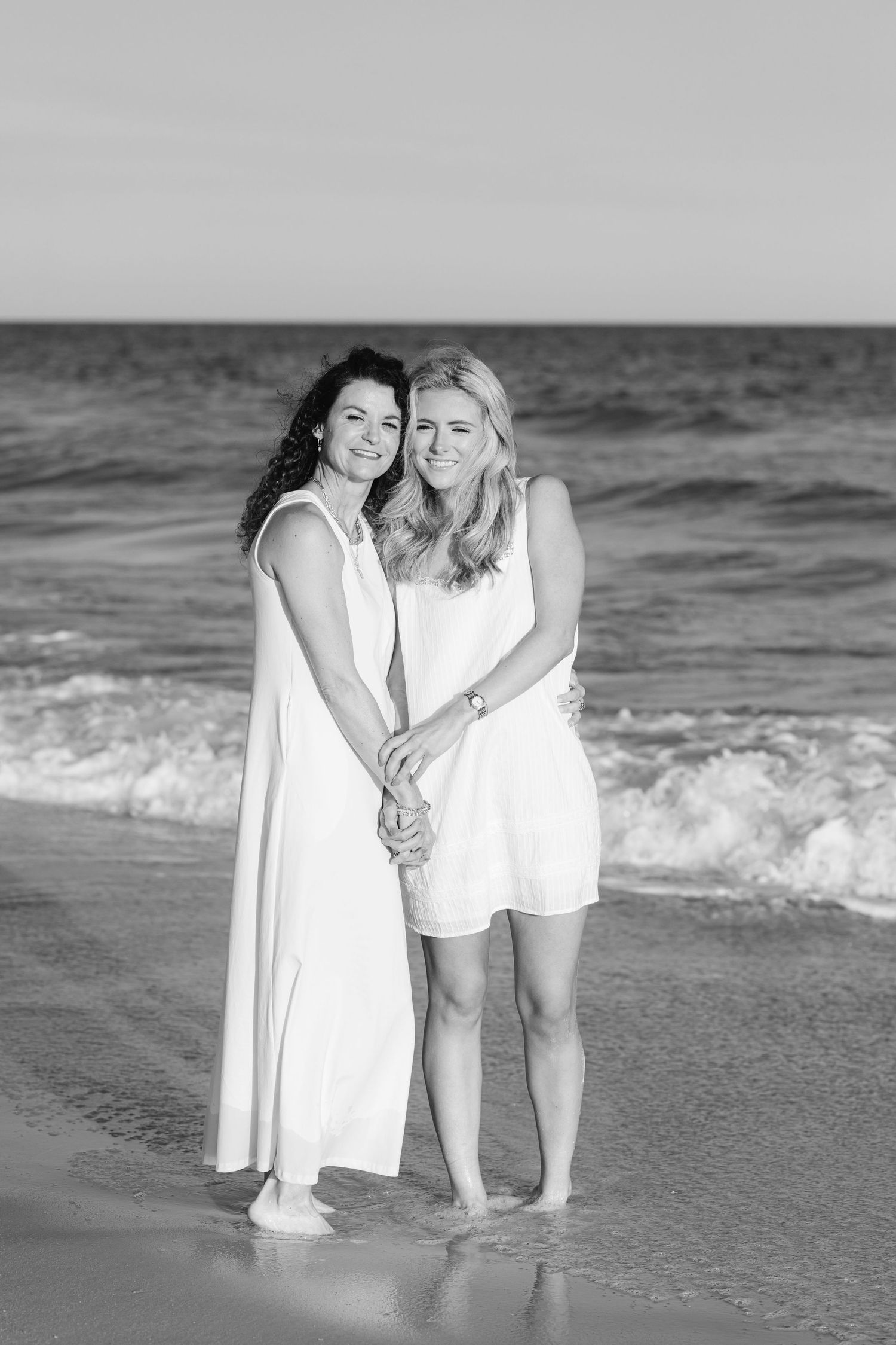 Black and white photo of two women in white dresses holding hands and smiling by ocean waves.