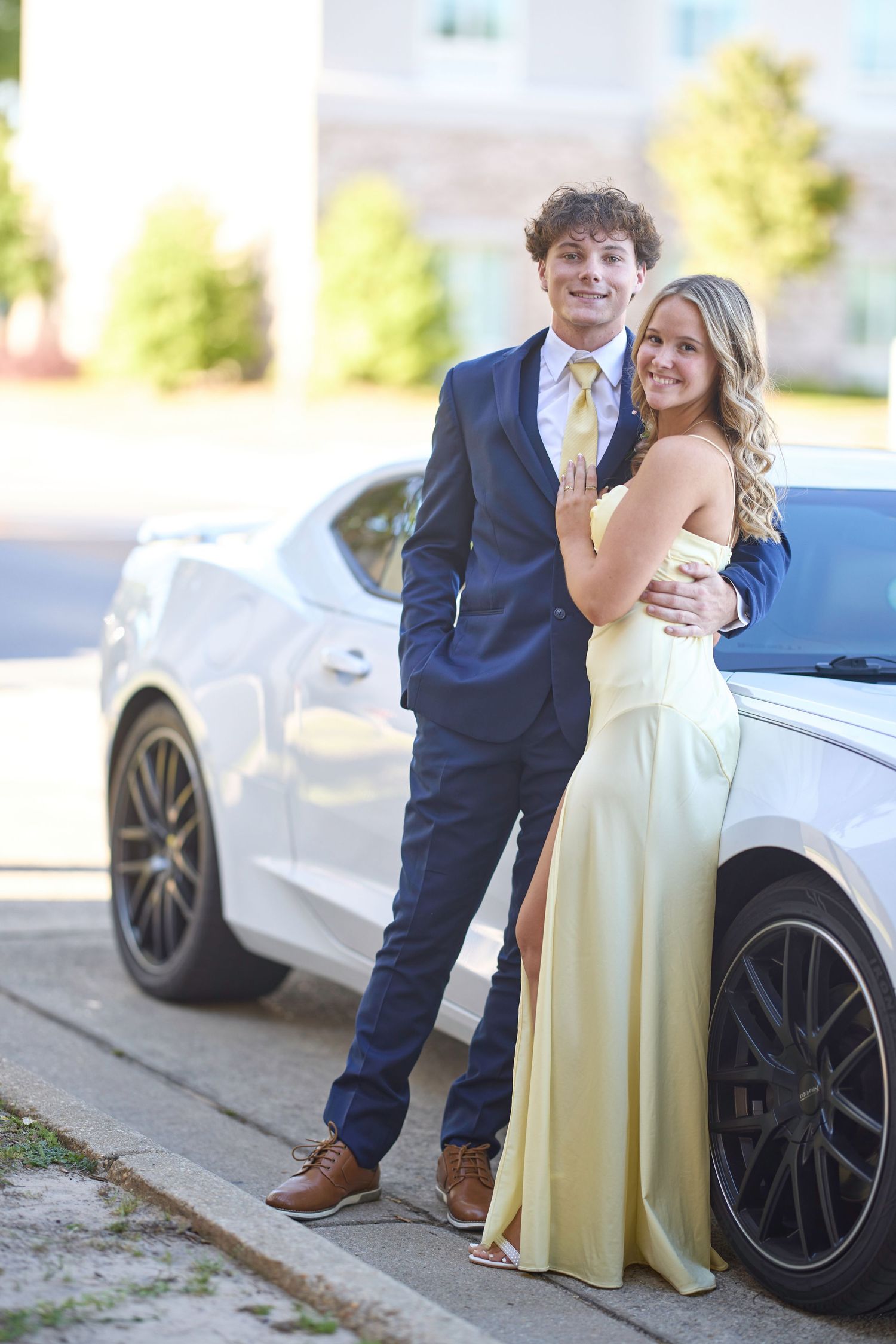 A couple poses together outdoors in formal attire next to a white luxury car.