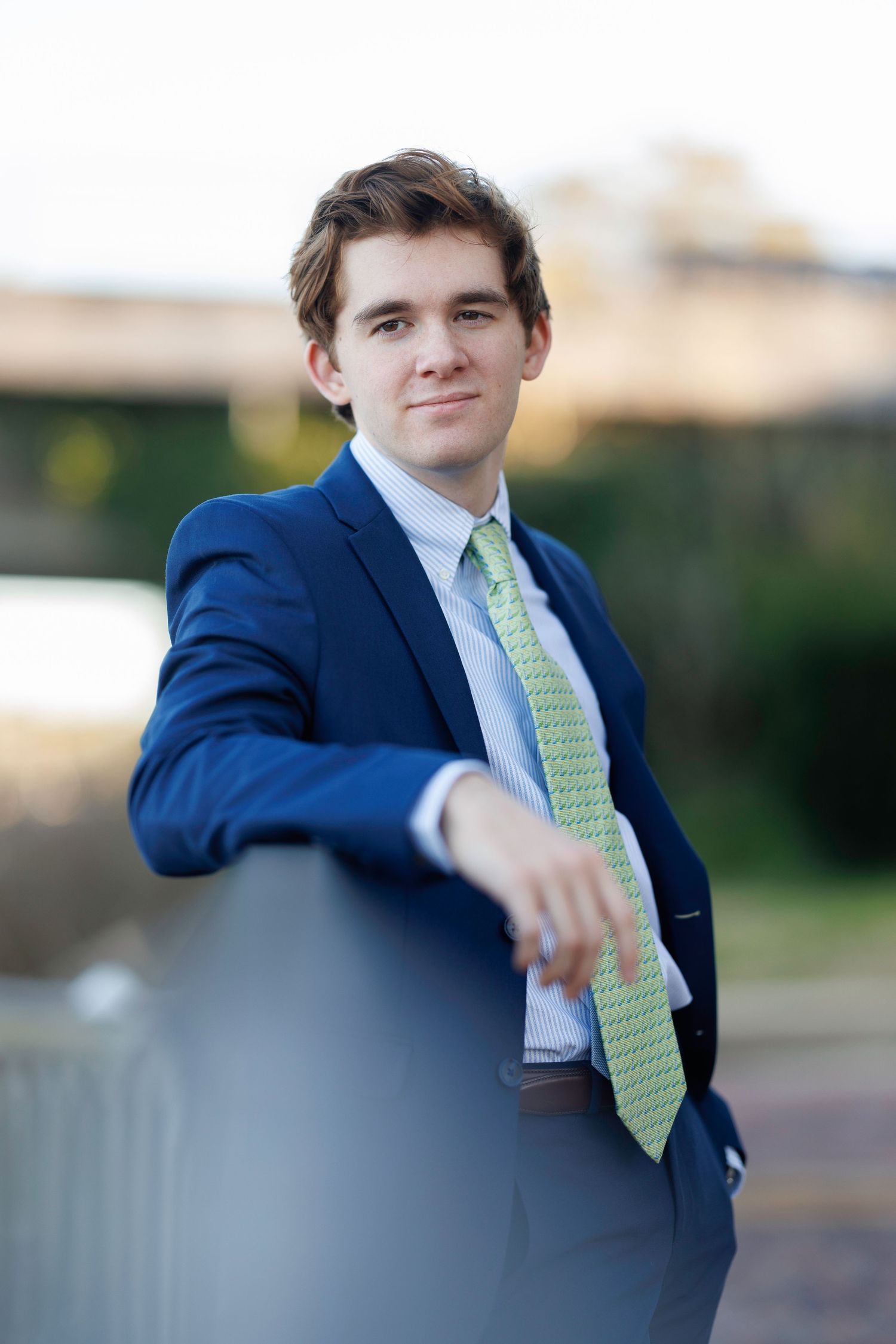 Young professional man in navy suit and green tie smiling outdoors, leaning on a railing.