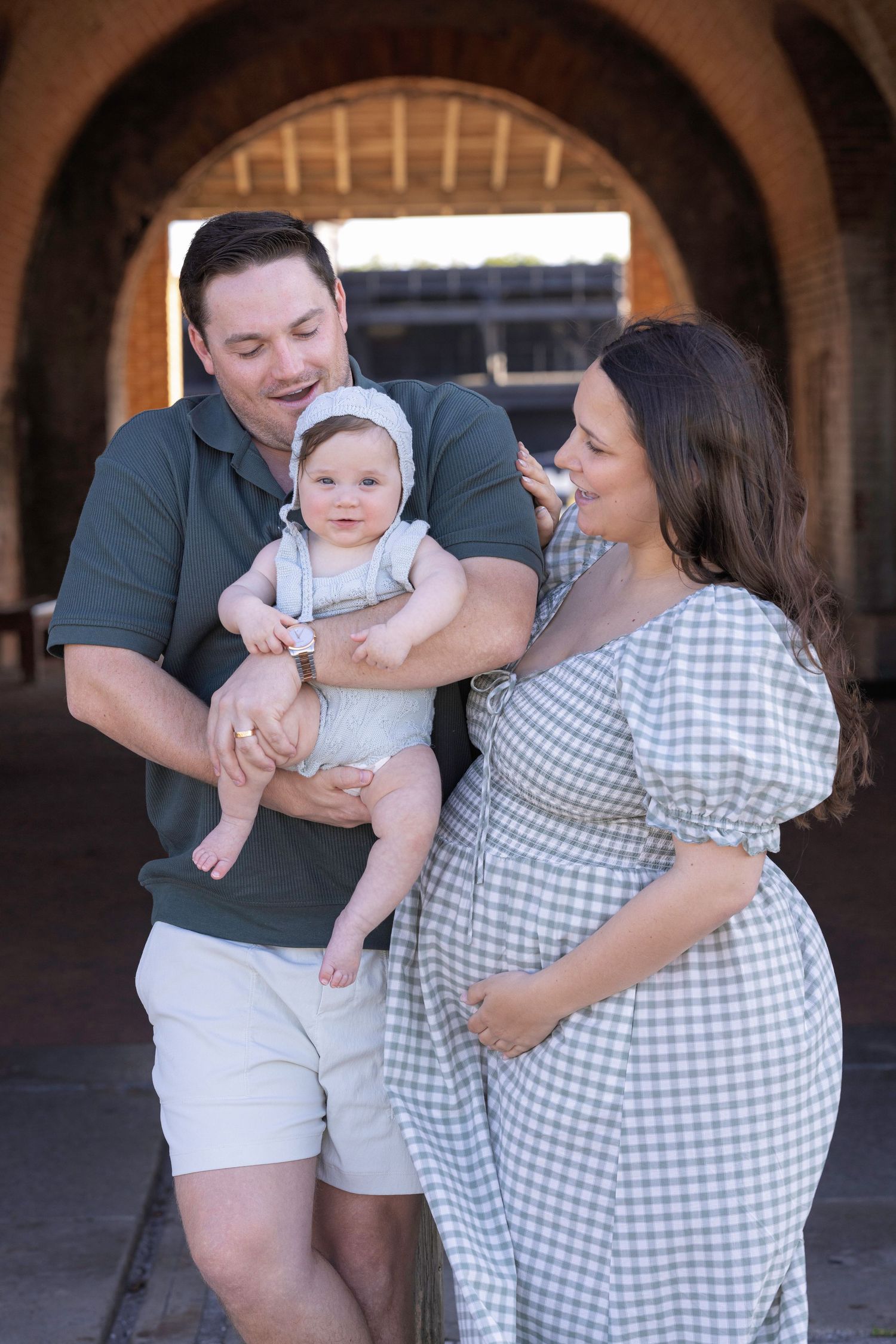 Happy family with man holding baby and pregnant woman in gingham dress posing under brick archway outdoors.