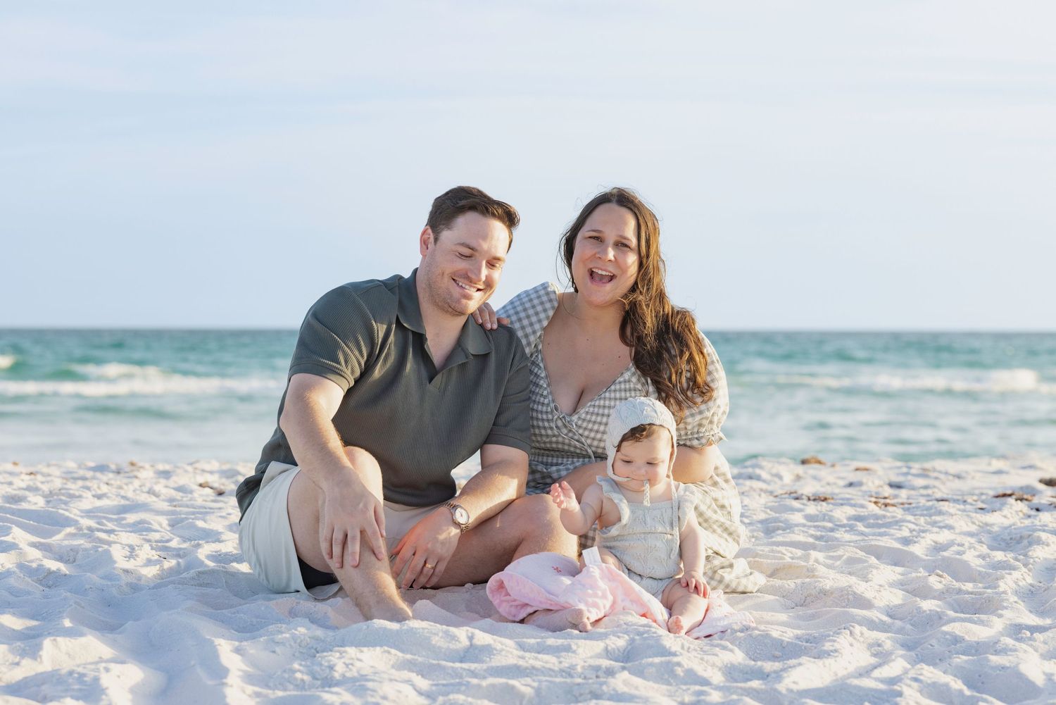 Family of three smiling on white sand beach, parents sitting with baby in pink outfit, ocean waves in background.