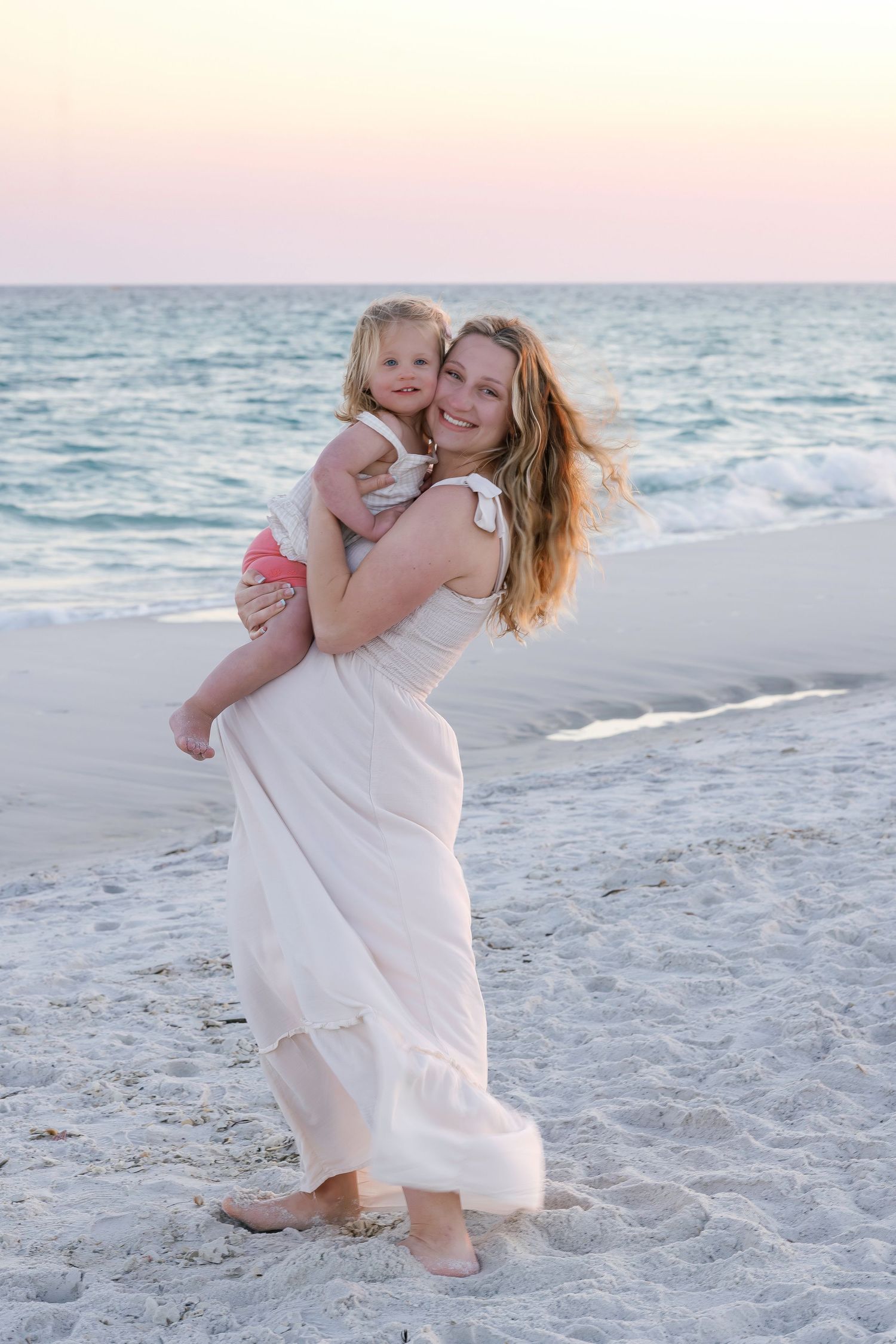 A smiling mother in a white maxi dress holds her young daughter on a beach at sunset.