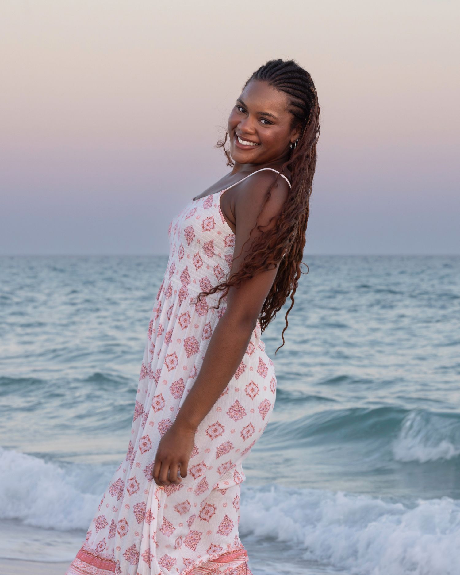 Smiling woman in floral sundress with braids poses on beach at sunset with ocean waves behind her.