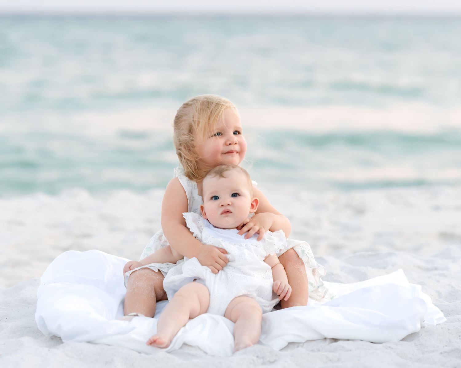 Two young sisters in white dresses cuddle on a white sandy beach with turquoise ocean waves in the background.